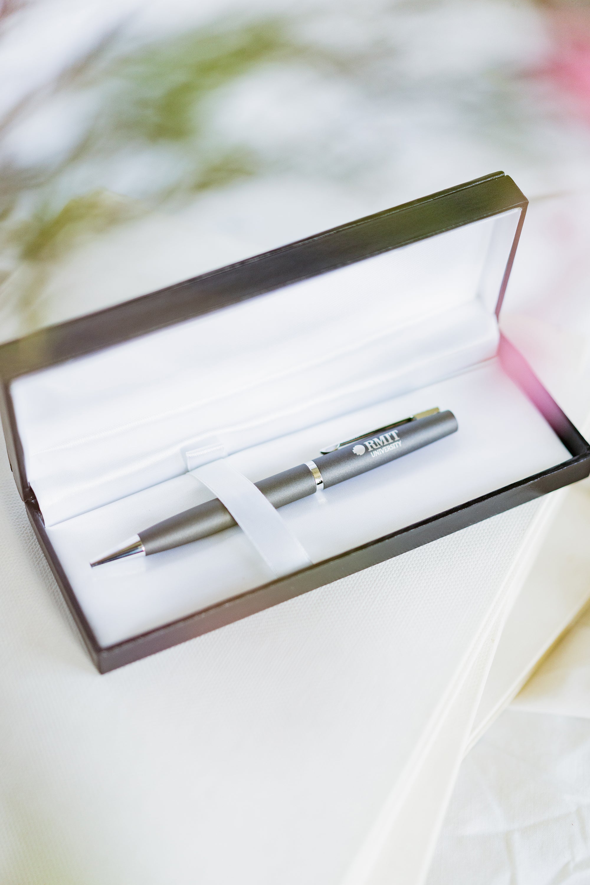 Corporate pen with a sleek charcoal finish displayed inside a black gift box with a soft silver interior lining. The pen features a polished metallic clip and tip, with “RMIT University” engraved near the clip. The gift box lid is open, showing the pen resting securely inside. The product is photographed on a light white tabletop for the RMIT Store.