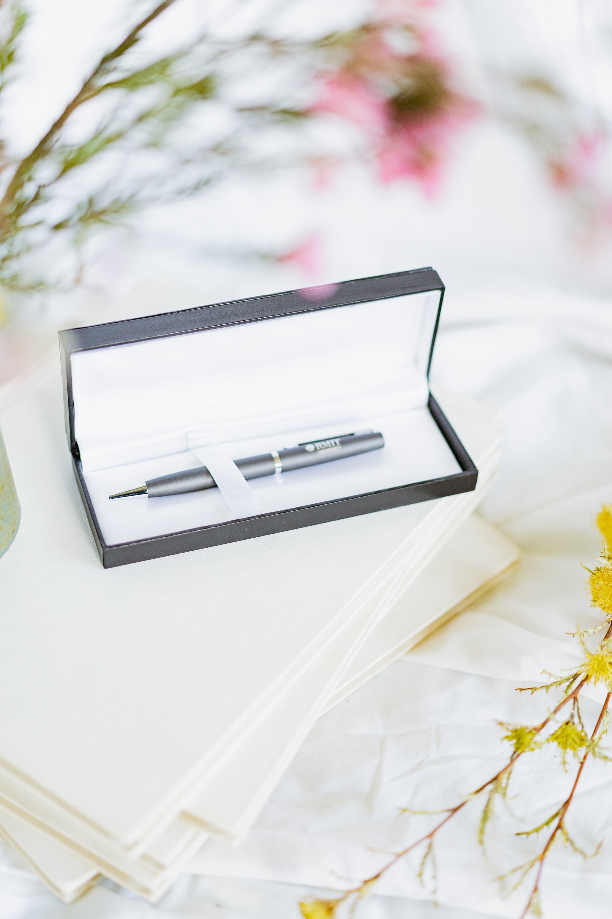Corporate pen with a sleek charcoal finish displayed inside a black gift box with a soft silver interior lining. The pen features a polished metallic clip and tip, with “RMIT University” engraved near the clip. The open gift box is placed on a light white tabletop, shown from a wider angle that includes more of the surrounding surface. The product is presented for the RMIT Store.