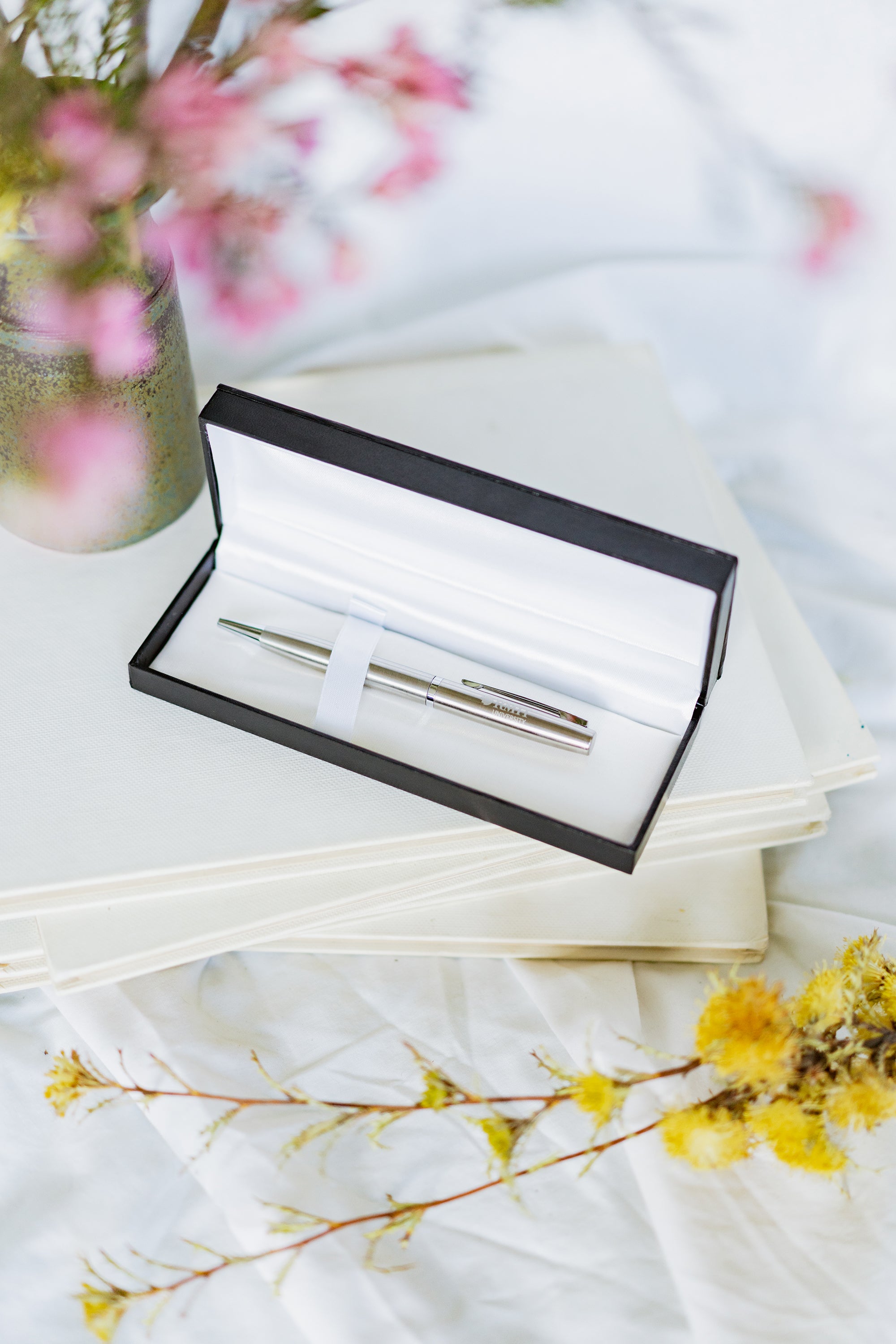 Corporate pen with a polished silver finish resting diagonally on a stack of hardcover books with plain spines showing no visible text. The pen features a metallic clip and tip with “RMIT University” engraved near the clip. Pink flowers are arranged to the left of the books, and the scene is set on a light white tabletop for the RMIT Store.
