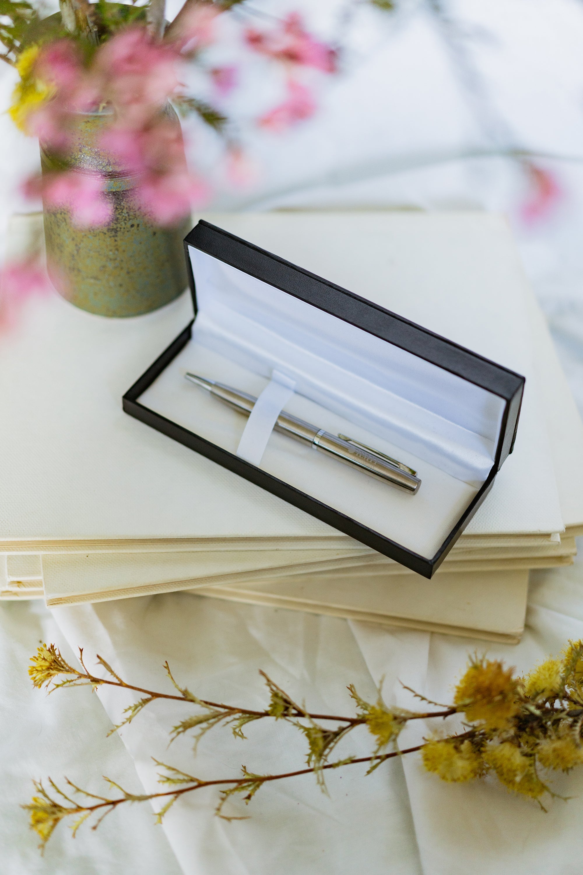 Corporate pen with a polished silver finish resting diagonally on a stack of hardcover books. The pen features a metallic clip and tip with “RMIT University” engraved near the clip. Pink flowers are arranged to the left of the books, and the scene is set on a light white tabletop for the RMIT Store.