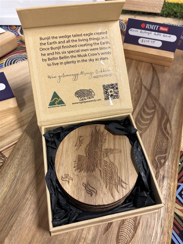 Creator Story Series Round Coasters Set of 8 displayed inside an open branded gift box with black tissue paper at RMIT Store, showing a stack of round wooden coasters with a laser-engraved wedge-tailed eagle design and surrounding symbolic motifs, with printed storytelling text and logos visible on the inside lid including Ngarga Warendj branding, Australian Made mark and QR code.