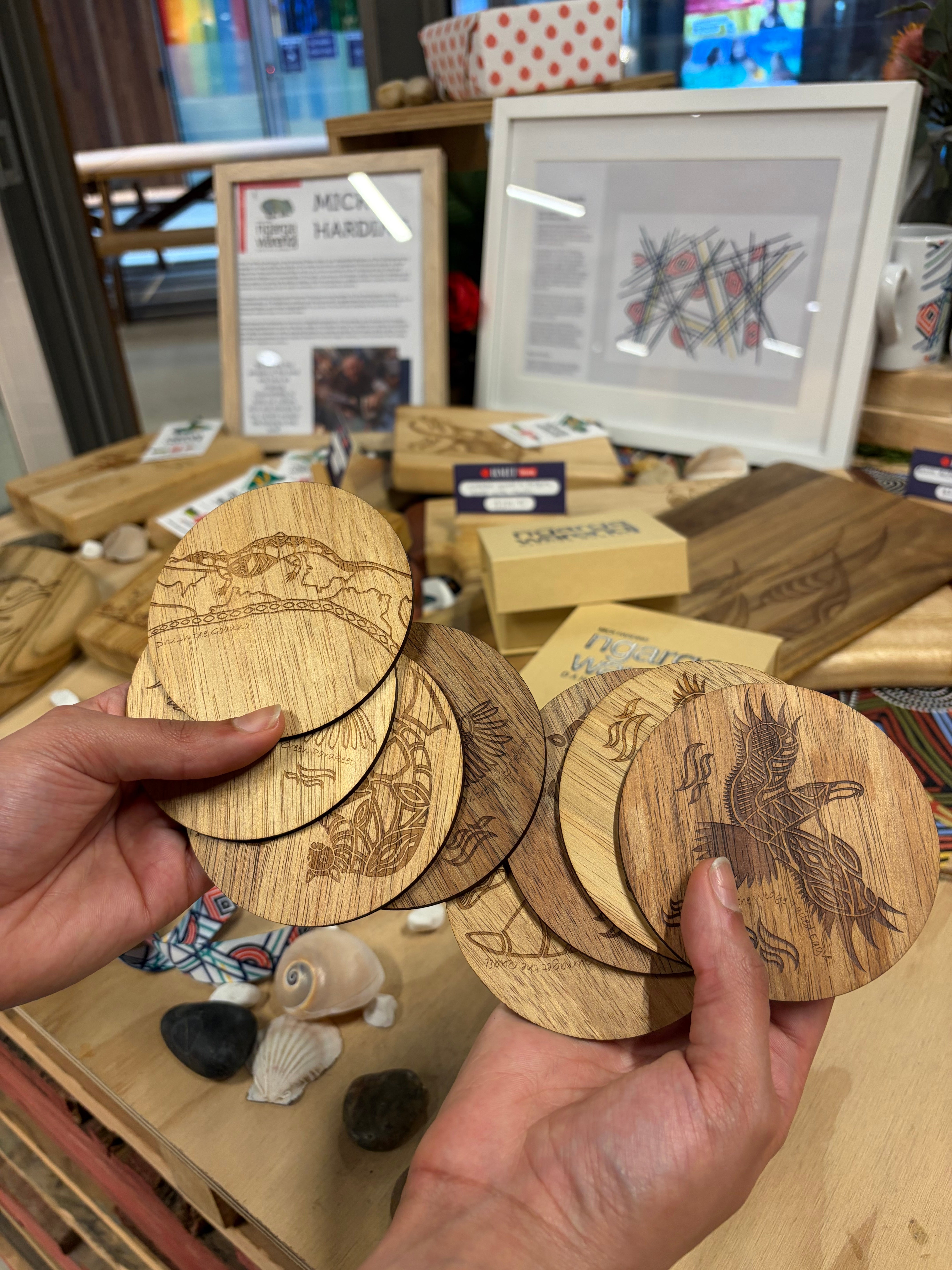 Creator Story Series Round Coasters Set of 8 made from light-toned wood with detailed laser-engraved Indigenous illustrations, held and fanned out in a model’s hands inside the RMIT Store, showing multiple distinct artwork designs including animals, landscape elements and symbolic linework, with fine etched detailing visible on each coaster surface and wooden product displays and framed artwork in the background.