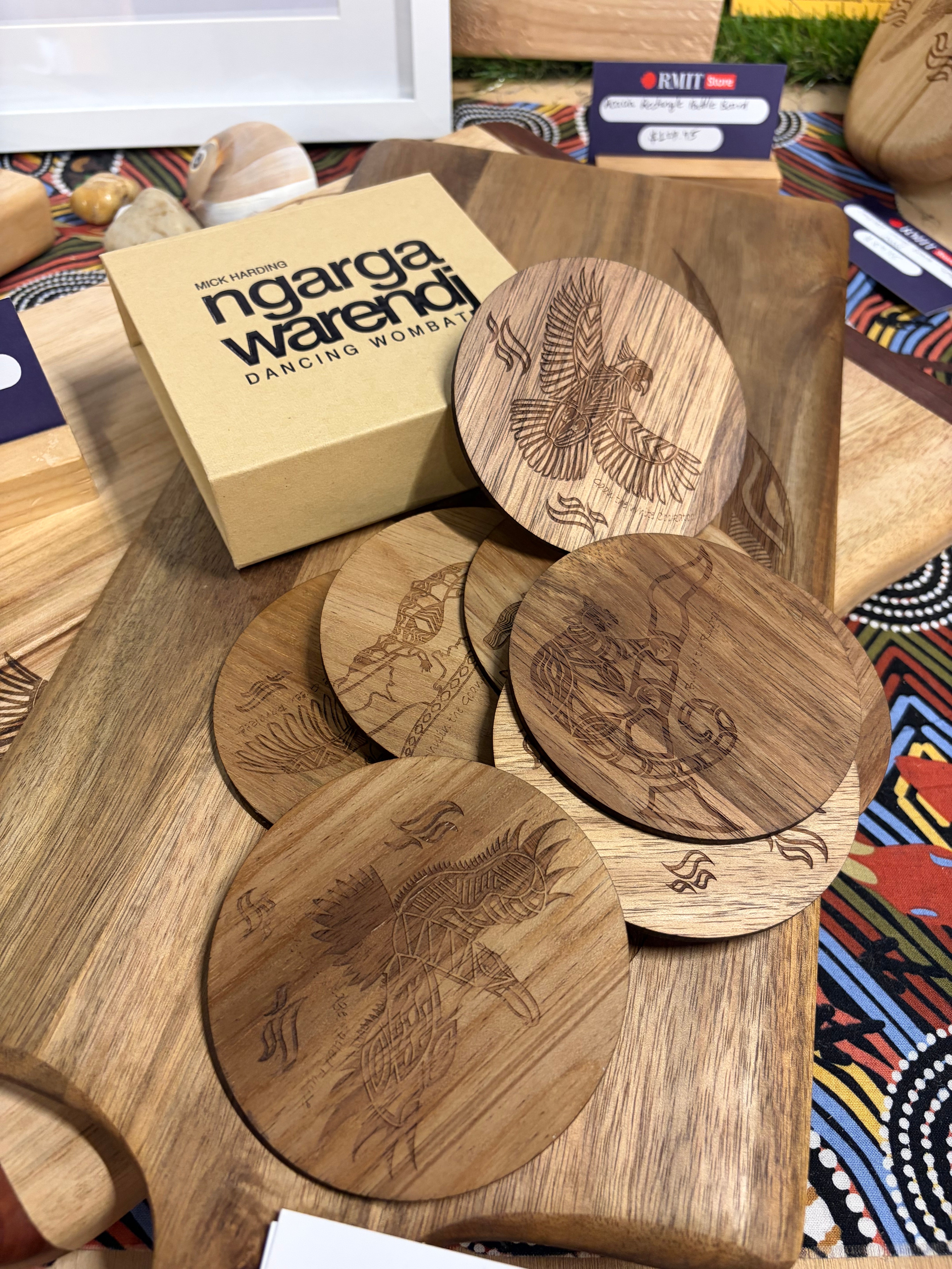 Creator Story Series Round Coasters Set of 8 displayed on a wooden serving board beside a beige Ngarga Warendj Dancing Wombat gift box at RMIT Store, showing layered light and dark wood round coasters with intricate laser-engraved Indigenous artwork including flying birds, animals, and symbolic patterns, with fine etched signatures and detailing visible on the surface and surrounding in-store cultural merchandise in the background.