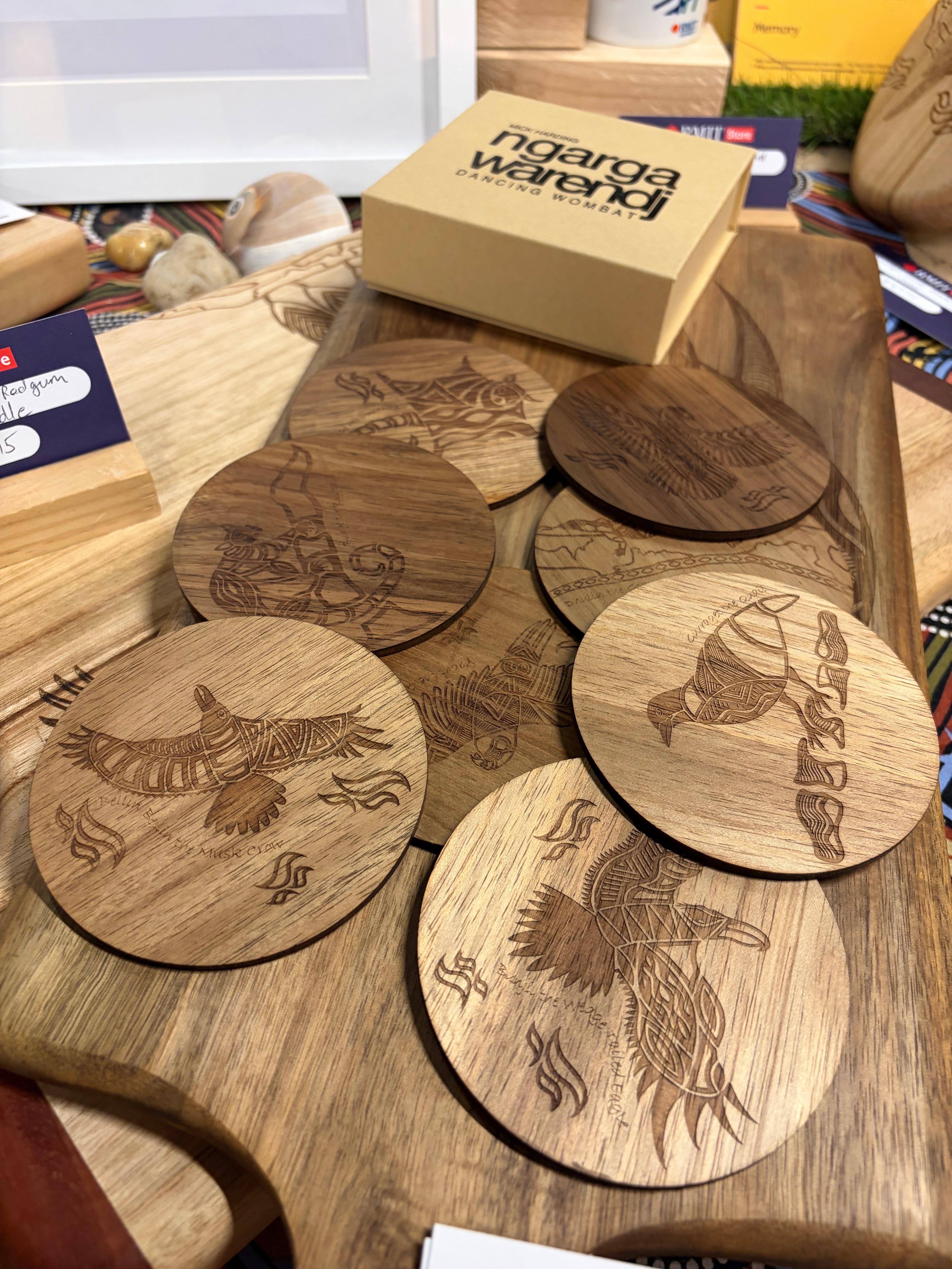 Creator Story Series Round Coasters Set of 8 featuring light and medium-toned wooden round coasters with detailed laser-engraved Indigenous bird and symbolic linework designs, arranged and overlapping on a large timber serving board in-store at RMIT Store, showing multiple distinct artworks including a wedge-tailed eagle and landscape motifs, with visible fine etched detailing, natural wood grain, and a branded Ngarga Warendj gift box positioned behind the display.