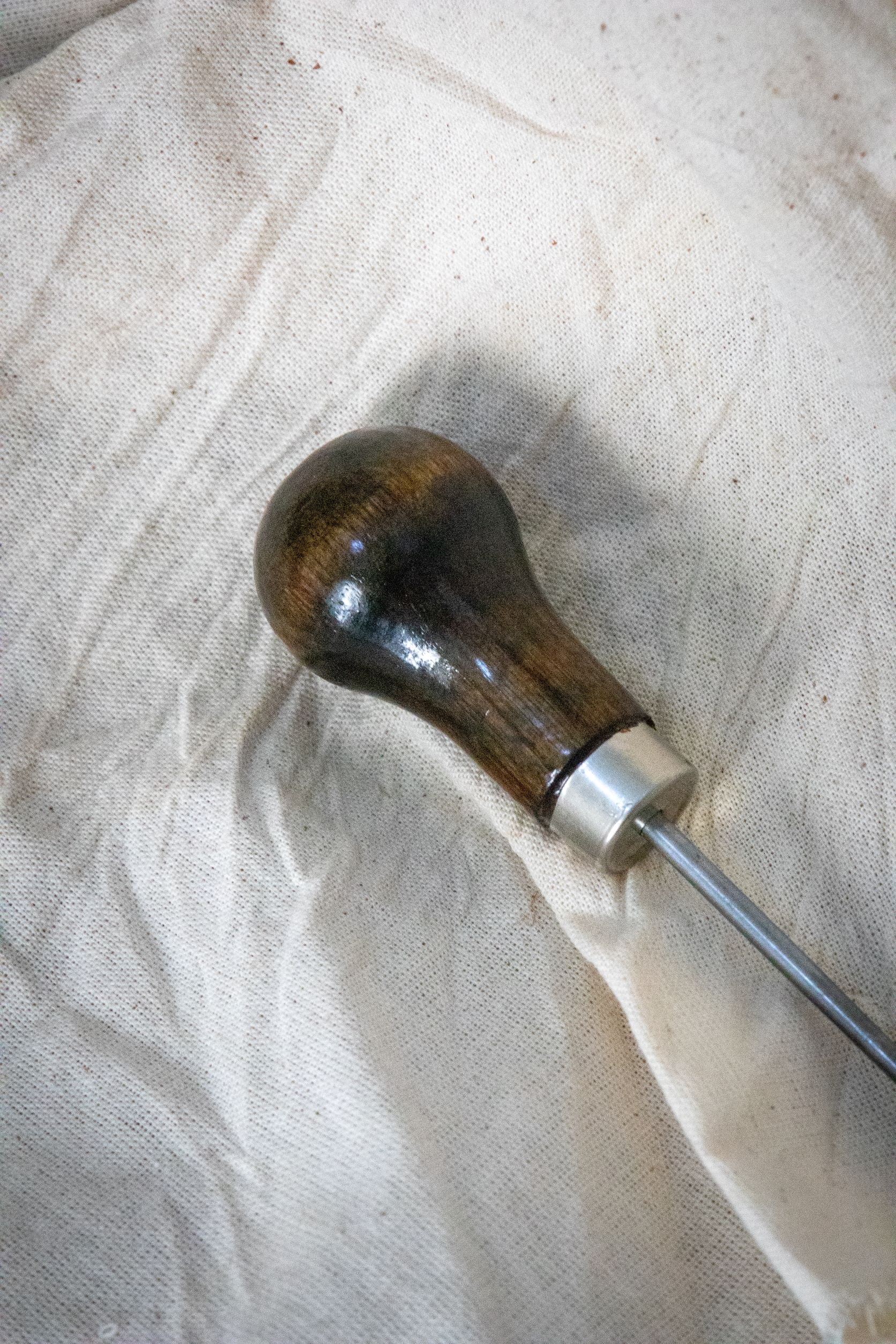 Close-up of the RMIT Store Dart Marker Tailors Awl handle, featuring a smooth, dark-stained wooden grip with a glossy finish and stainless steel collar, placed on a textured cream fabric background.