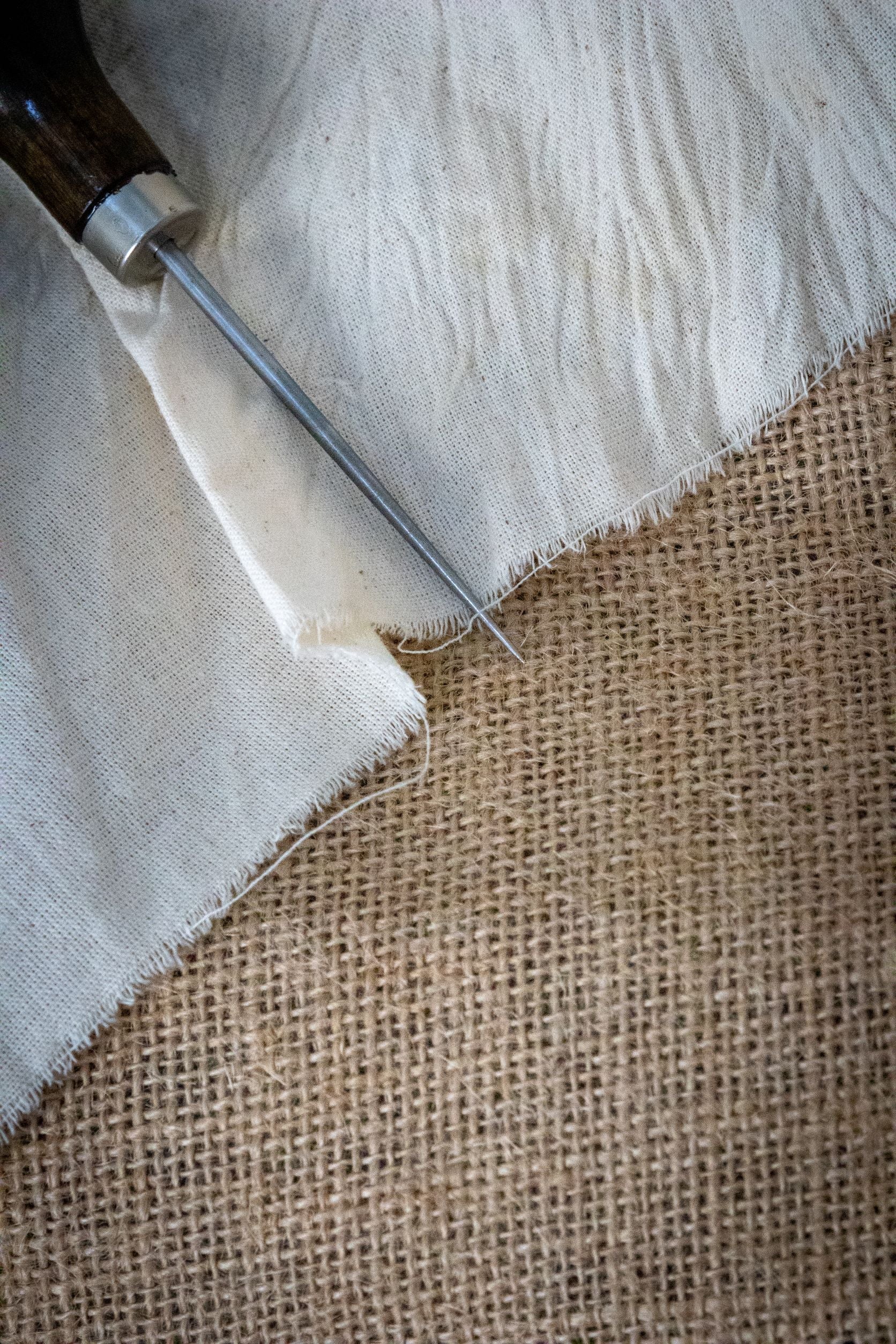 RMIT Store Dart Marker Tailors Awl shown from the side with the sharp steel point extended over both white cotton and coarse brown hessian fabric, highlighting the precision tip and material contrast.