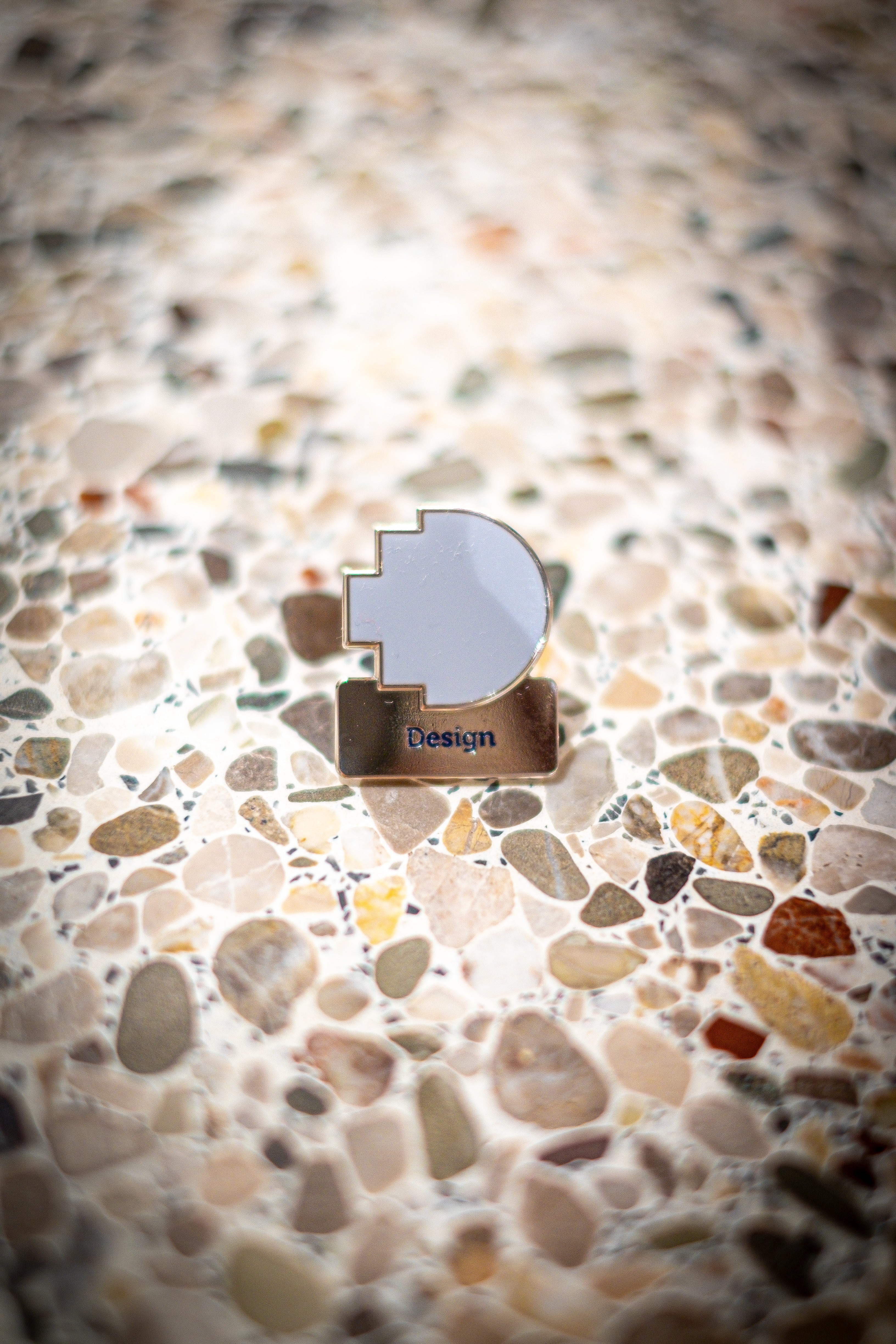 Front view of the Design Discipline pin placed on a light marble background, featuring the RMIT pixel logo in light blue above a silver square with the word "Design". Available at the RMIT Store.