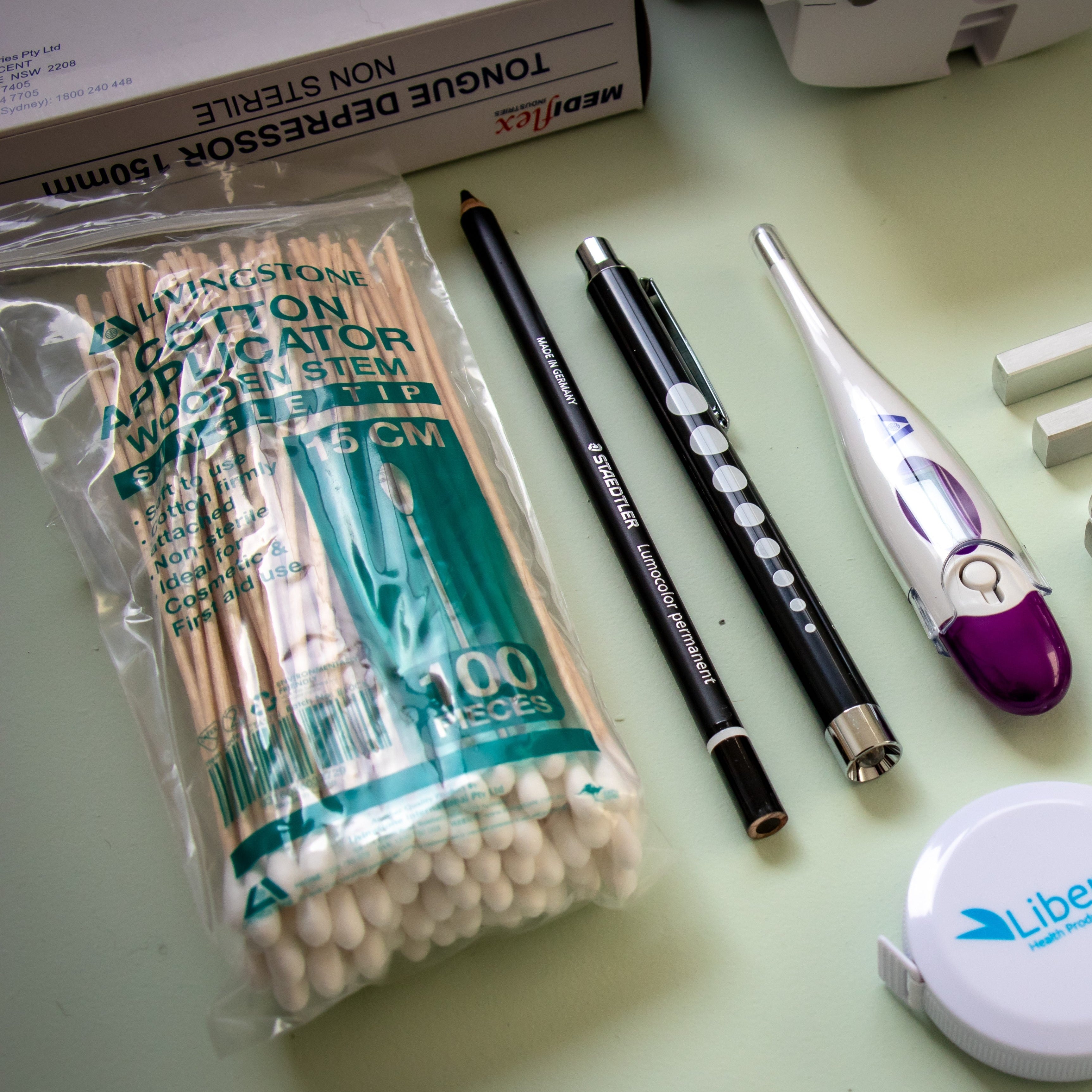 Assorted medical diagnostic tools from the RMIT Store displayed on a table. Includes cotton applicators, a Glasochrom permanent marker, a Liberty penlight torch, and a digital clinical thermometer, emphasising their utility for various clinical applications.