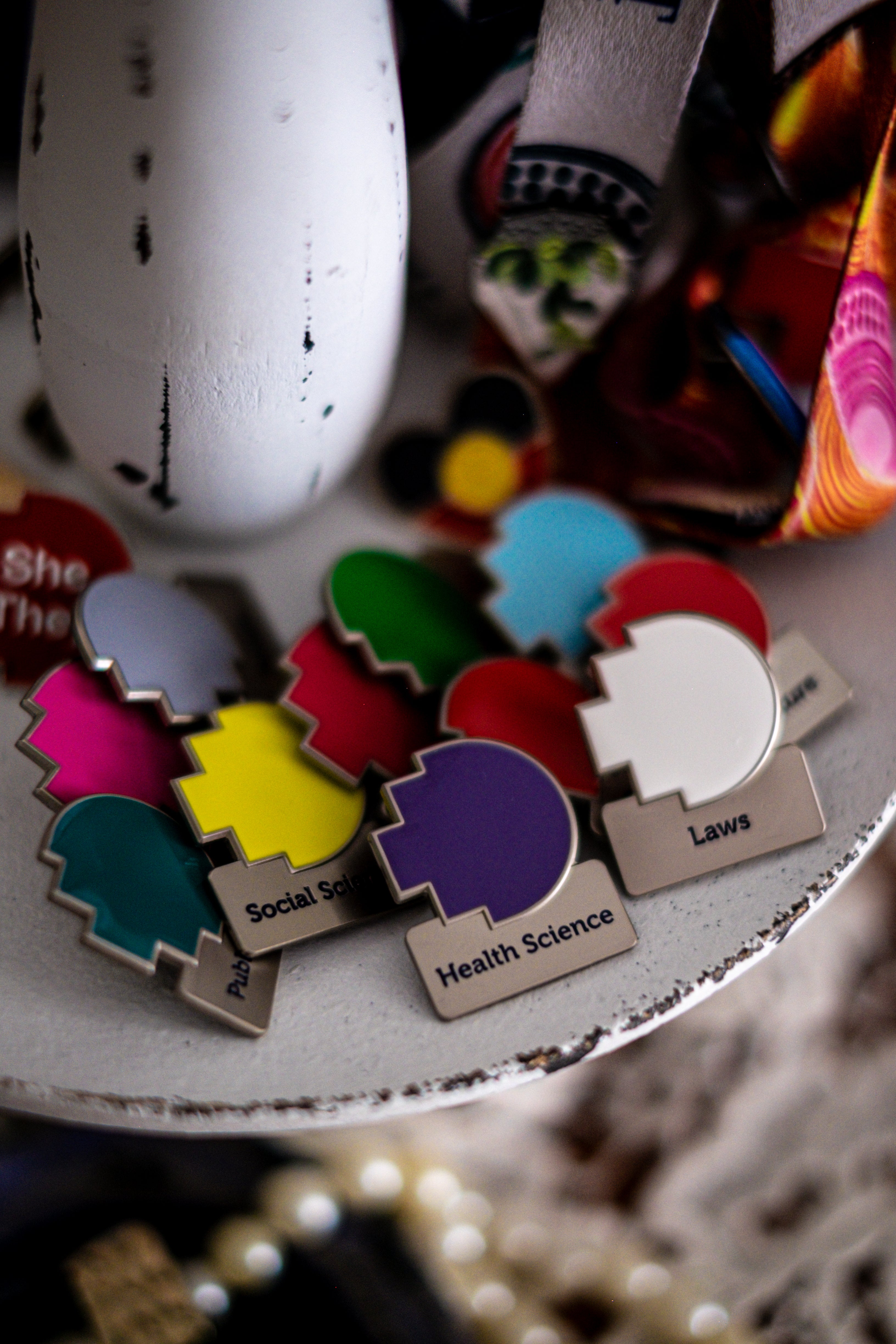 Assorted RMIT Discipline Lapel Matte Pins in multiple colours and fields, arranged overlapping on a distressed white circular stand beside a lanyard and a pronoun pin, with pearls and lace underneath. Available at the RMIT Store.
