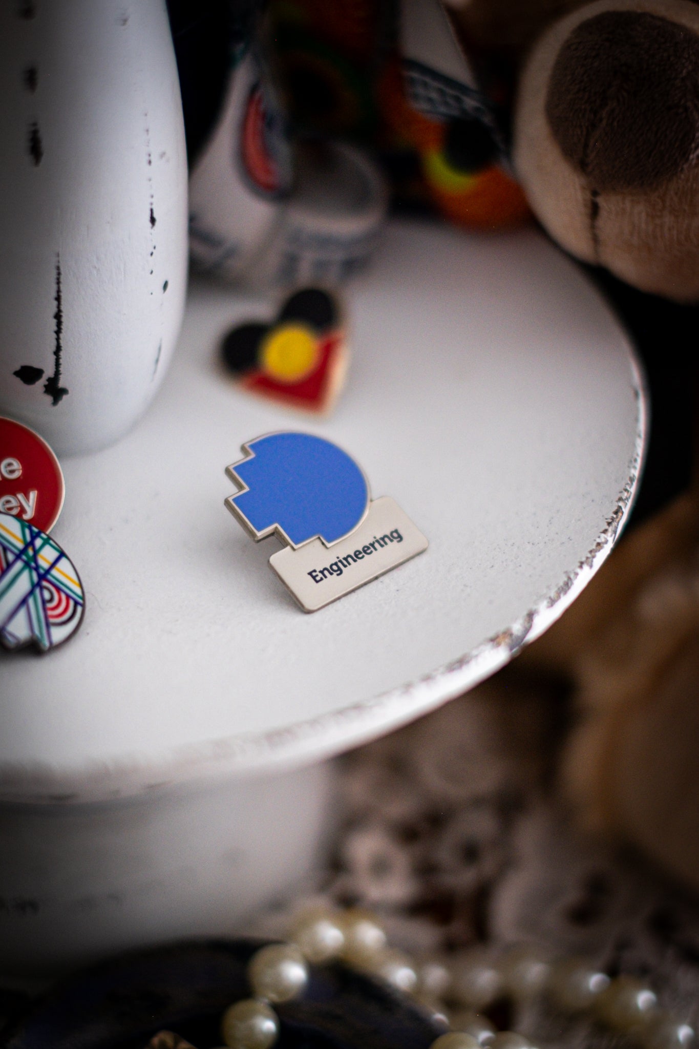 RMIT Discipline Lapel Matte Pin in medium blue for Engineering, displayed upright on a distressed white circular stand beside other enamel pins, a lanyard and an RMIT Graduation Bear, with pearls arranged at the base. Available at the RMIT Store.