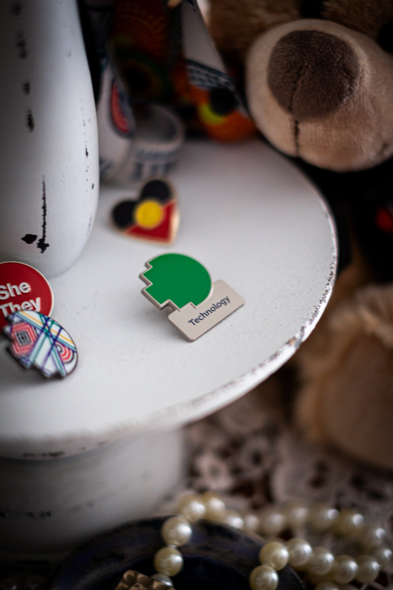 RMIT Discipline Lapel Matte Pin in green for Technology, displayed upright on a distressed white circular stand beside other enamel pins, a lanyard and an RMIT Graduation Bear, with pearls arranged at the base. Available at the RMIT Store.