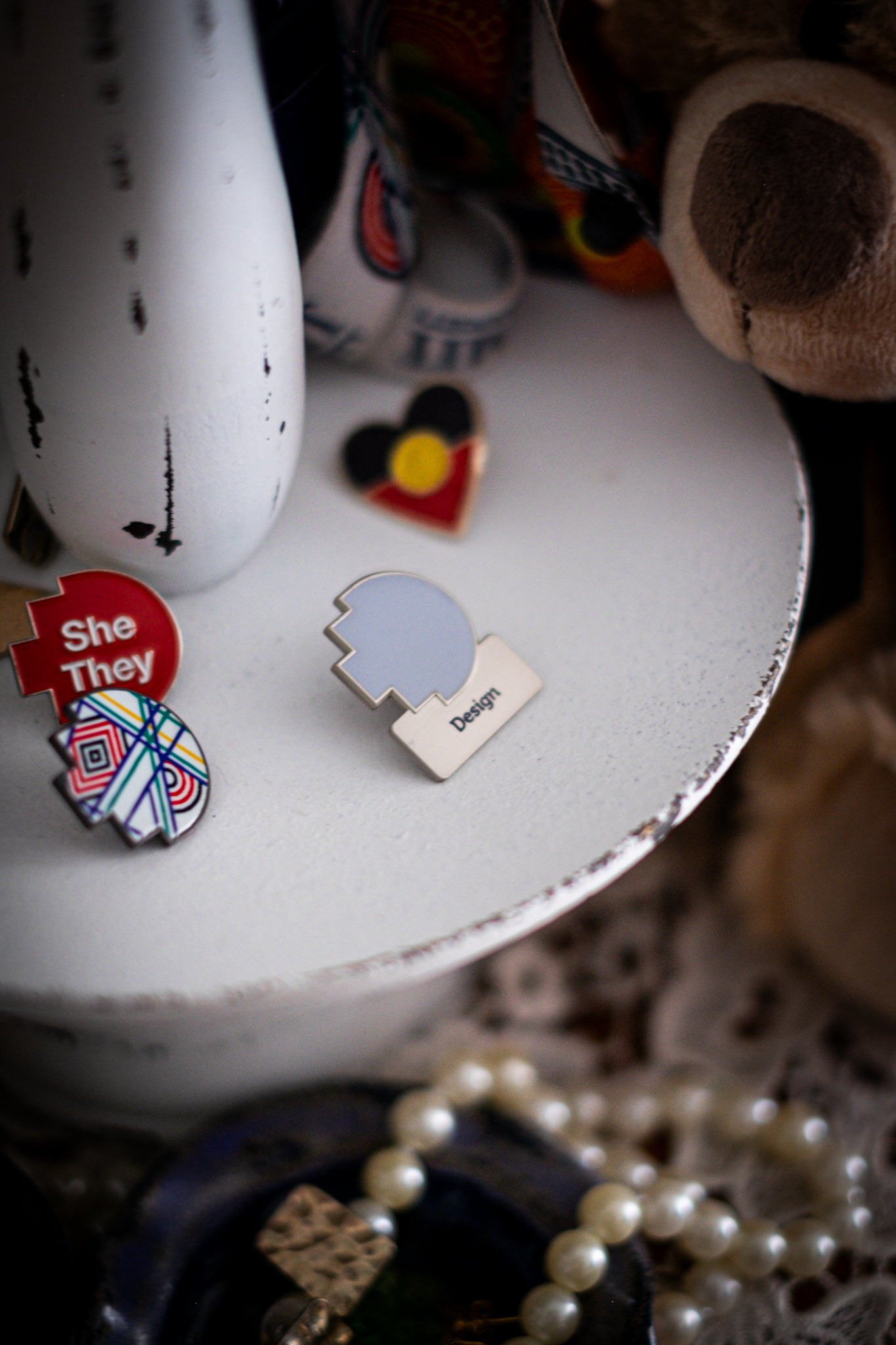 RMIT Discipline Lapel Matte Pin in grey for Design, displayed upright on a distressed white circular stand beside other enamel pins, a lanyard, and an RMIT Graduation Bear, with pearls arranged at the base. Available at the RMIT Store.