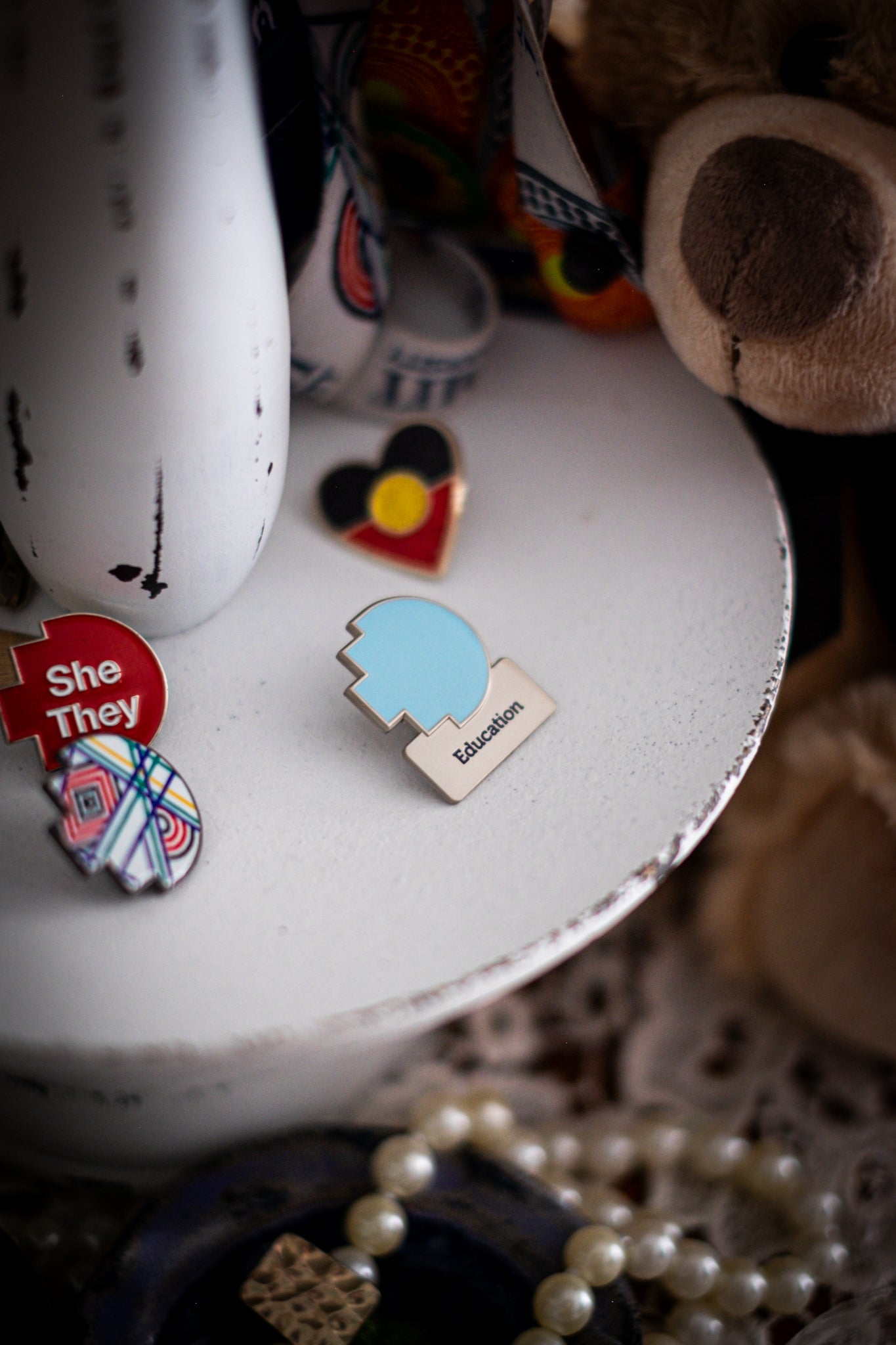 RMIT Discipline Lapel Matte Pin in light blue for Education, displayed upright on a distressed white circular stand beside other enamel pins, a lanyard, and an RMIT Graduation Bear, with pearls arranged at the base. Available at the RMIT Store.