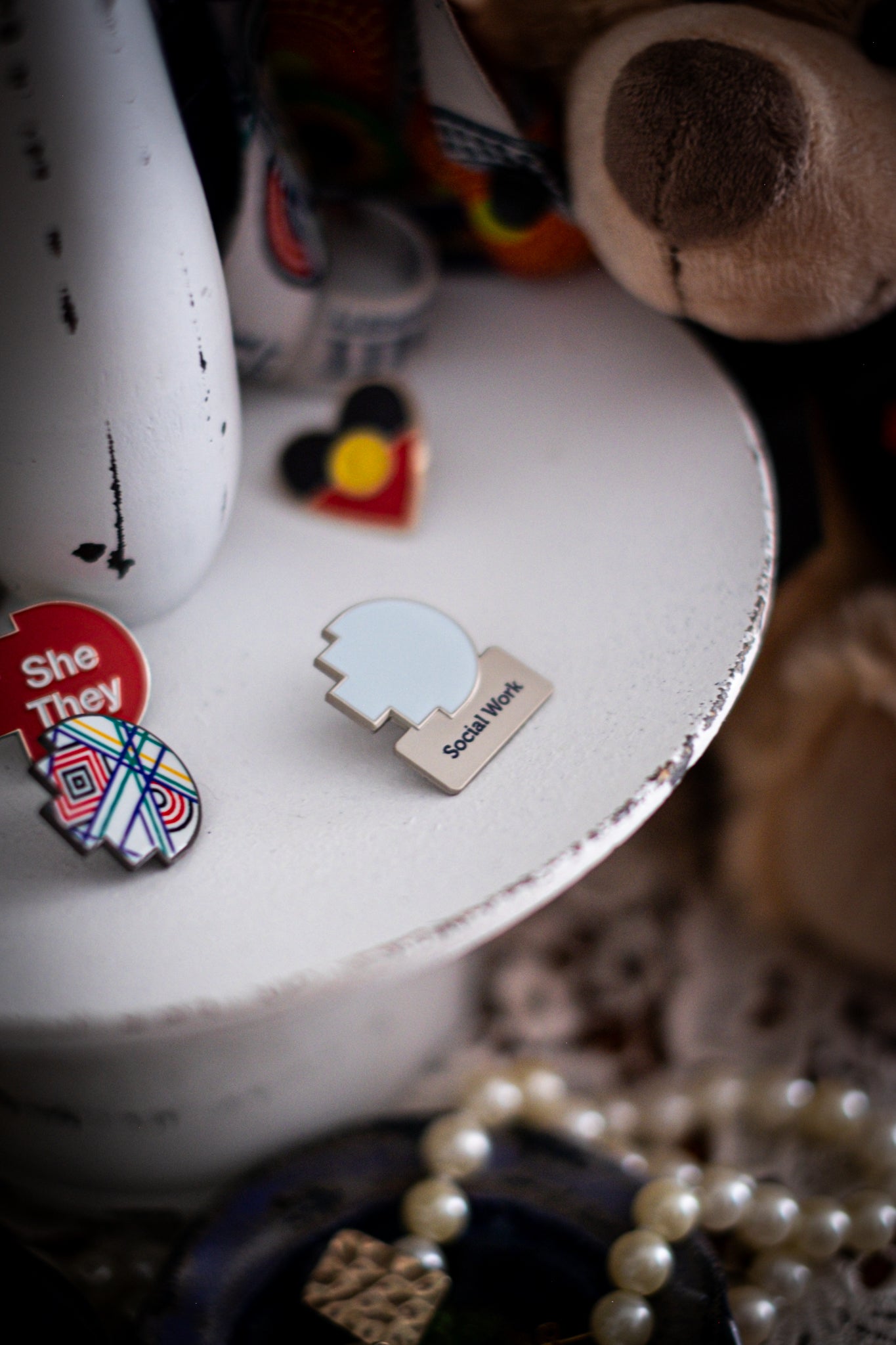 RMIT Discipline Lapel Matte Pin in light blue for Social Work, displayed upright on a distressed white circular stand beside other enamel pins, a lanyard and an RMIT Graduation Bear, with pearls arranged at the base. Available at the RMIT Store.