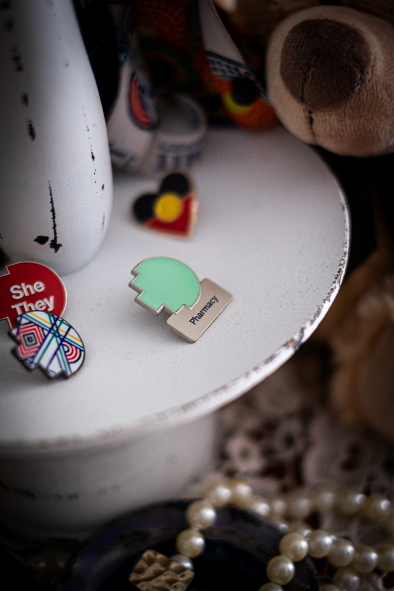 RMIT Discipline Lapel Matte Pin in mint green for Pharmacy, displayed upright on a distressed white circular stand beside other enamel pins, a lanyard and an RMIT Graduation Bear, with pearls arranged at the base. Available at the RMIT Store.