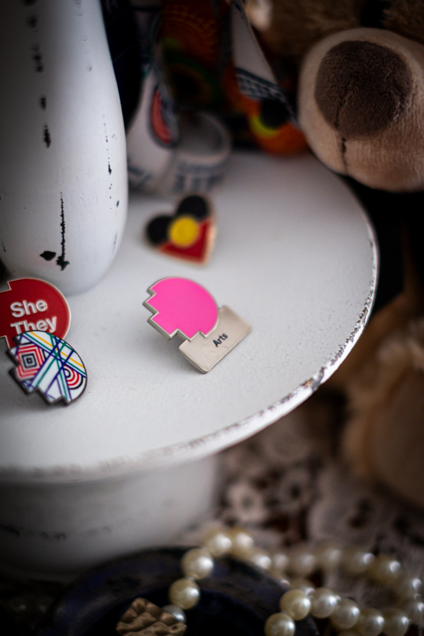 RMIT Discipline Lapel Matte Pin in bright pink for Arts, displayed upright on a distressed white circular stand beside other enamel pins, a lanyard and an RMIT Graduation Bear, with pearls arranged at the base. Available at the RMIT Store.