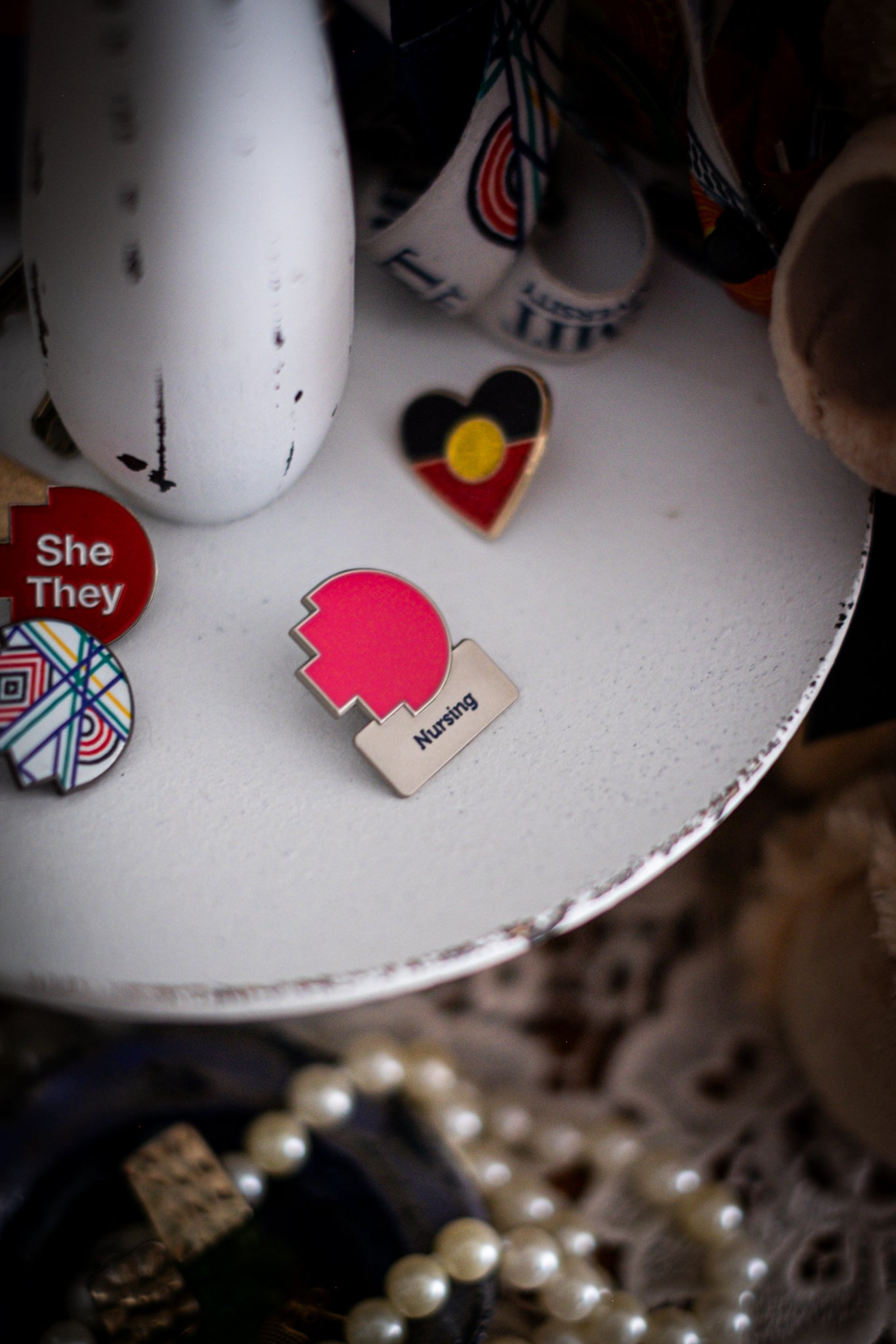 RMIT Discipline Lapel Matte Pin in pink for Nursing, displayed upright on a distressed white circular stand beside other enamel pins, a lanyard, and an RMIT Graduation Bear, with pearls arranged at the base. Available at the RMIT Store.