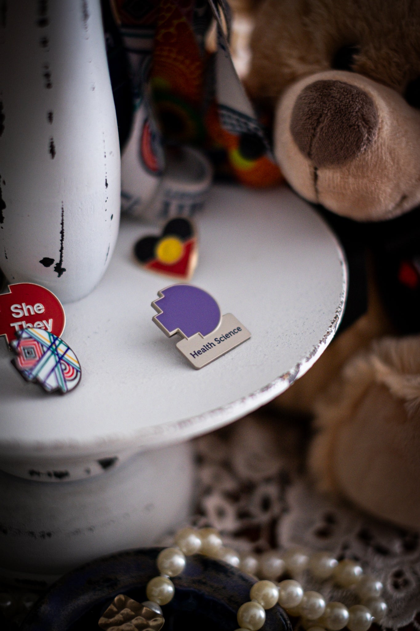 RMIT Discipline Lapel Matte Pin in lavender purple for Health Science, displayed upright on a distressed white circular stand beside other enamel pins, a lanyard and an RMIT Graduation Bear, with pearls arranged at the base. Available at the RMIT Store.