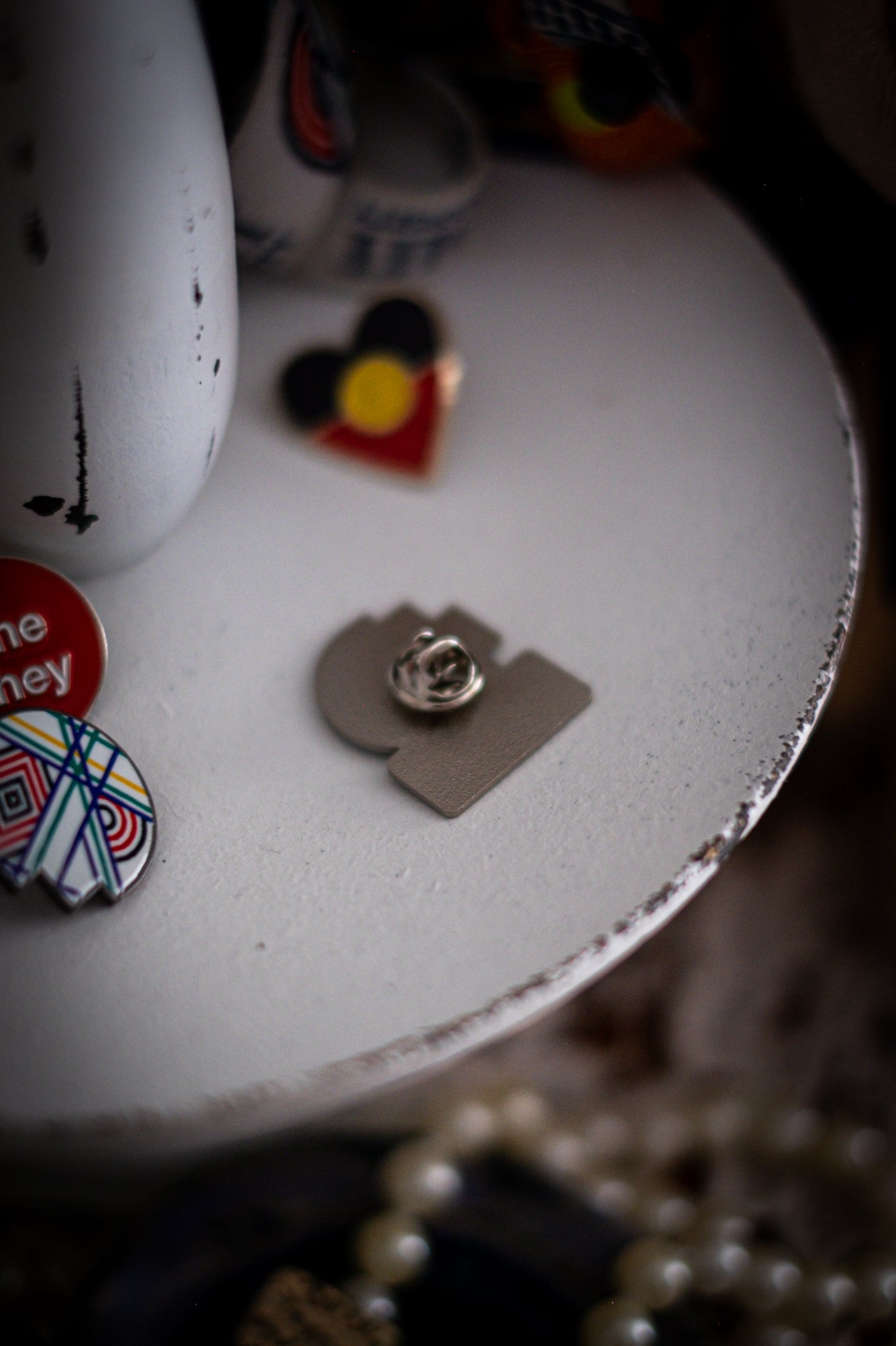 Rear view of RMIT Discipline Lapel Matte Pin showing matte silver metal finish with clasp fastening, displayed flat on a distressed white circular stand beside other enamel pins, a lanyard, and an RMIT Graduation Bear, with pearls arranged at the base. Available at the RMIT Store.
