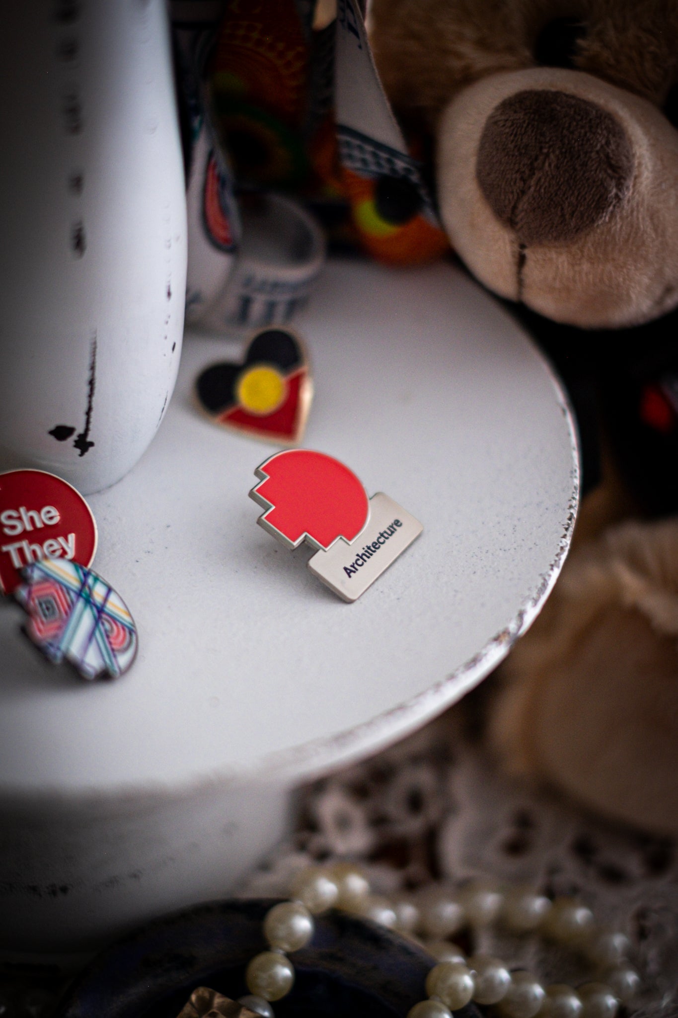 RMIT Discipline Lapel Matte Pin in red for Architecture, displayed upright on a distressed white circular stand beside other enamel pins, a lanyard, and an RMIT Graduation Bear, with pearls arranged at the base. Available at the RMIT Store.