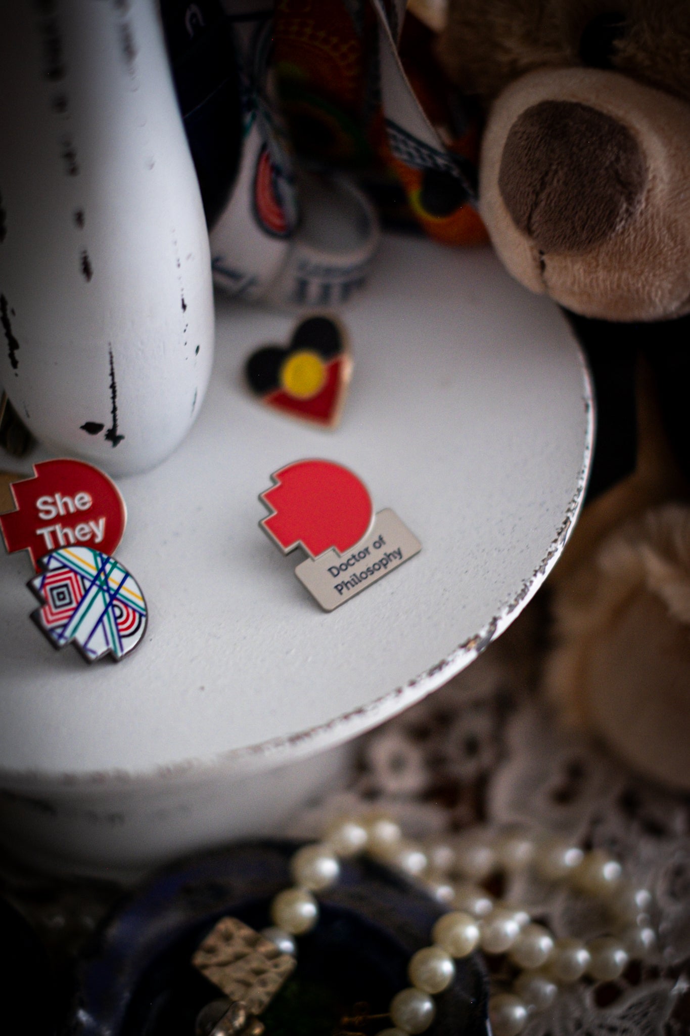 RMIT Discipline Lapel Matte Pin in red for Doctor of Philosophy, displayed upright on a distressed white circular stand beside other enamel pins, a lanyard, and an RMIT Graduation Bear, with pearls arranged at the base. Available at the RMIT Store.