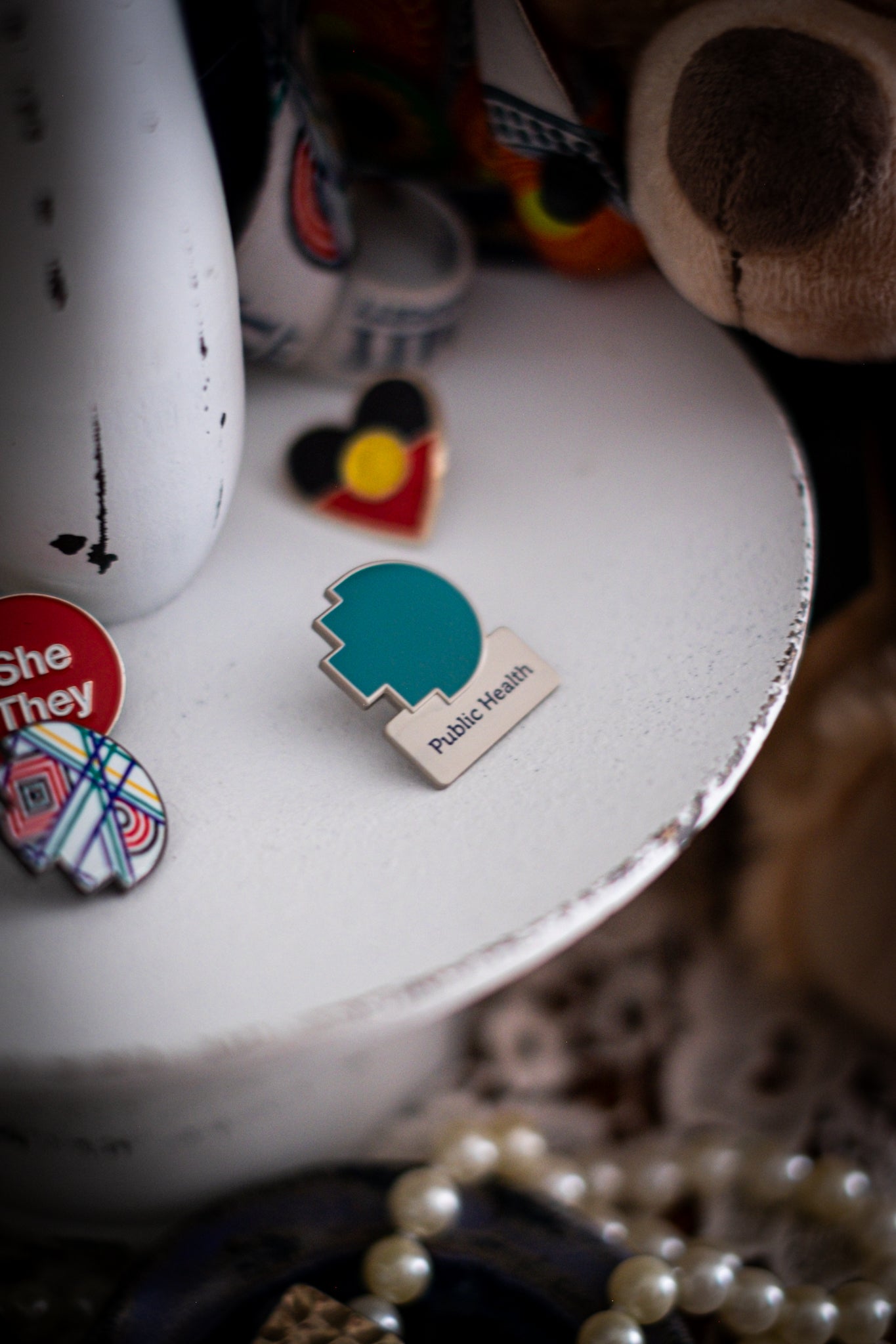 RMIT Discipline Lapel Matte Pin in teal blue for Public Health, displayed upright on a distressed white circular stand beside other enamel pins, a lanyard and an RMIT Graduation Bear, with pearls arranged at the base. Available at the RMIT Store.