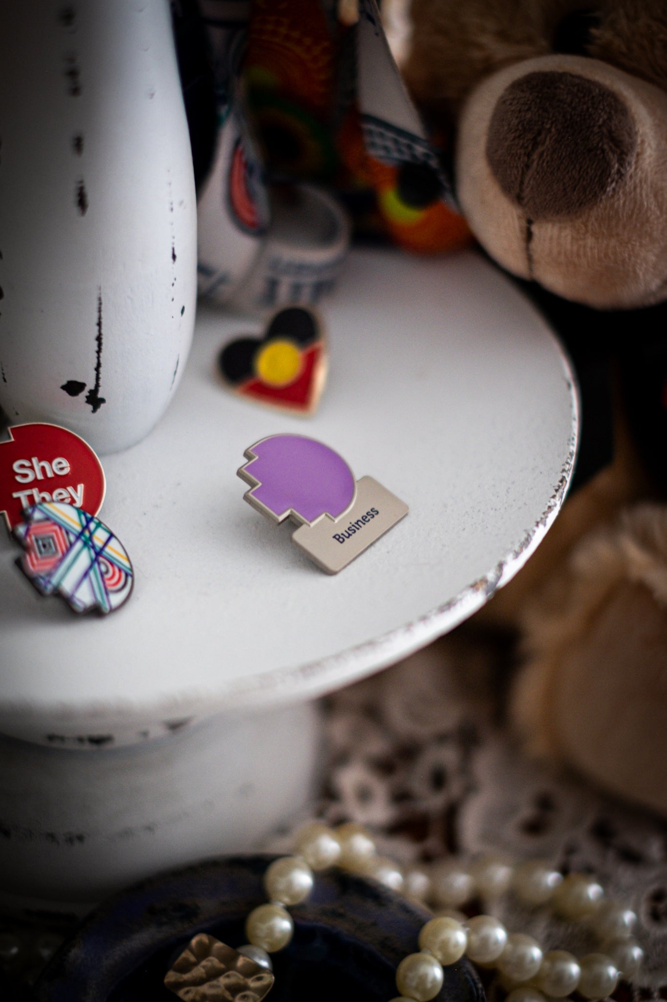 RMIT Discipline Lapel Matte Pin in light violet for Business, displayed upright on a distressed white circular stand beside other enamel pins, a lanyard and an RMIT Graduation Bear, with pearls arranged at the base. Available at the RMIT Store.