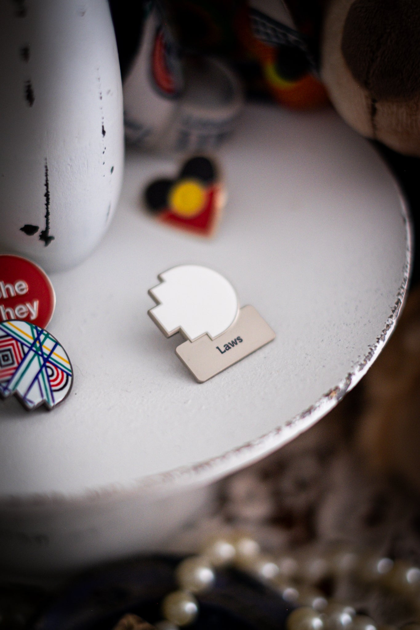 RMIT Discipline Lapel Matte Pin in white for Laws, displayed upright on a distressed white circular stand beside other enamel pins, a lanyard and an RMIT Graduation Bear, with pearls arranged at the base. Available at the RMIT Store.