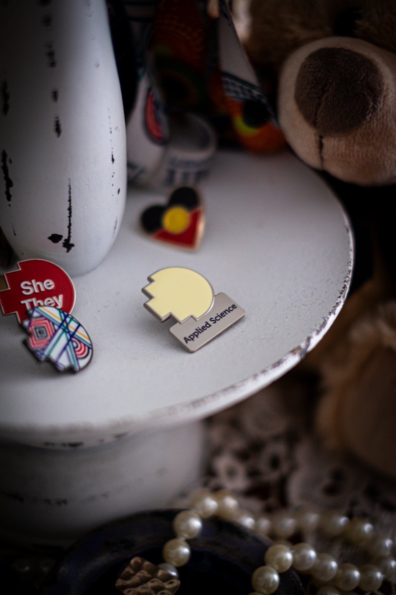 RMIT Discipline Lapel Matte Pin in pale yellow for Applied Science, displayed upright on a distressed white circular stand beside other enamel pins, a lanyard and an RMIT Graduation Bear, with pearls arranged at the base. Available at the RMIT Store.