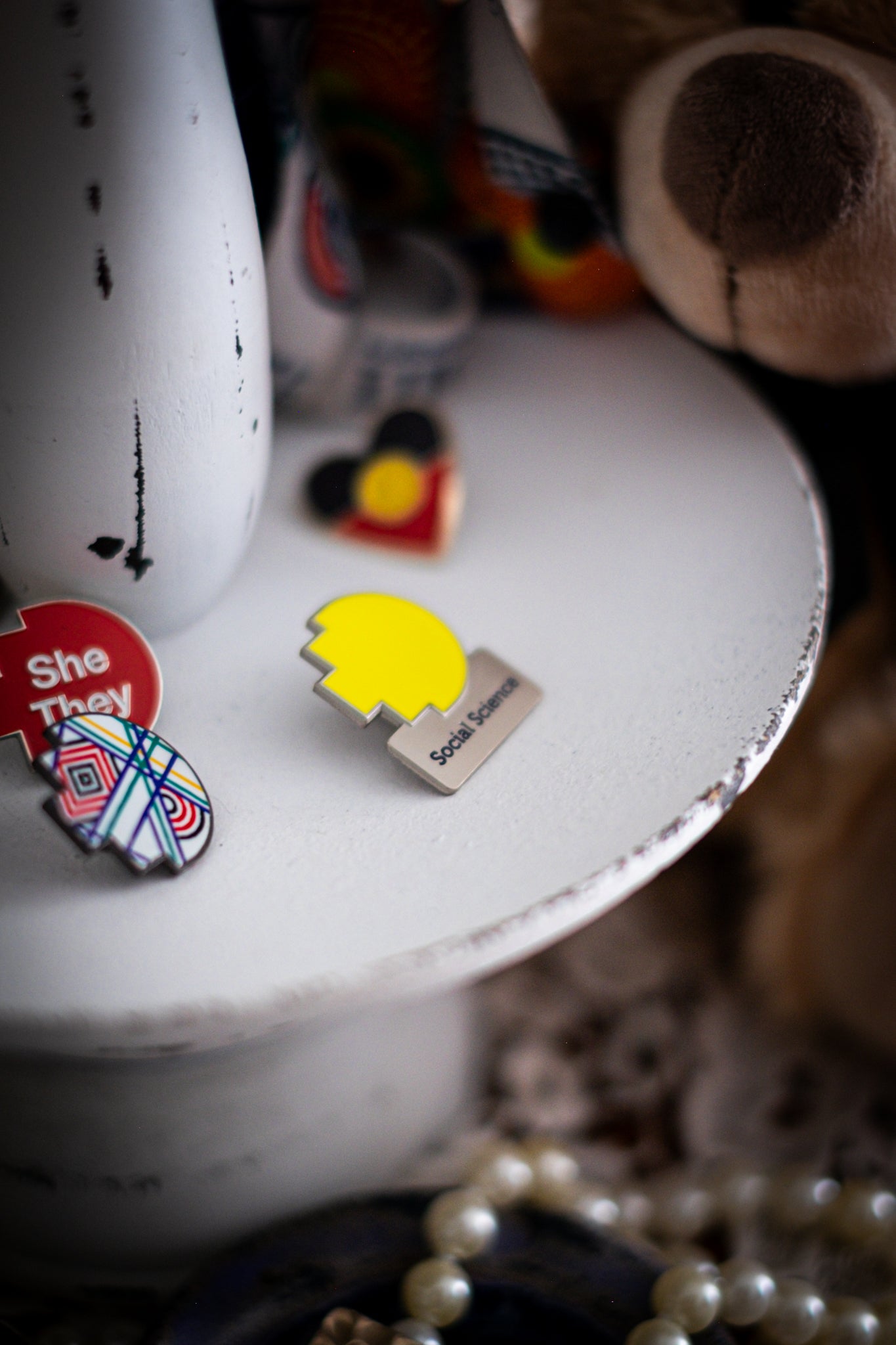 RMIT Discipline Lapel Matte Pin in yellow for Social Science, displayed upright on a distressed white circular stand beside other enamel pins, a lanyard, and an RMIT Graduation Bear, with pearls arranged at the base. Available at the RMIT Store.