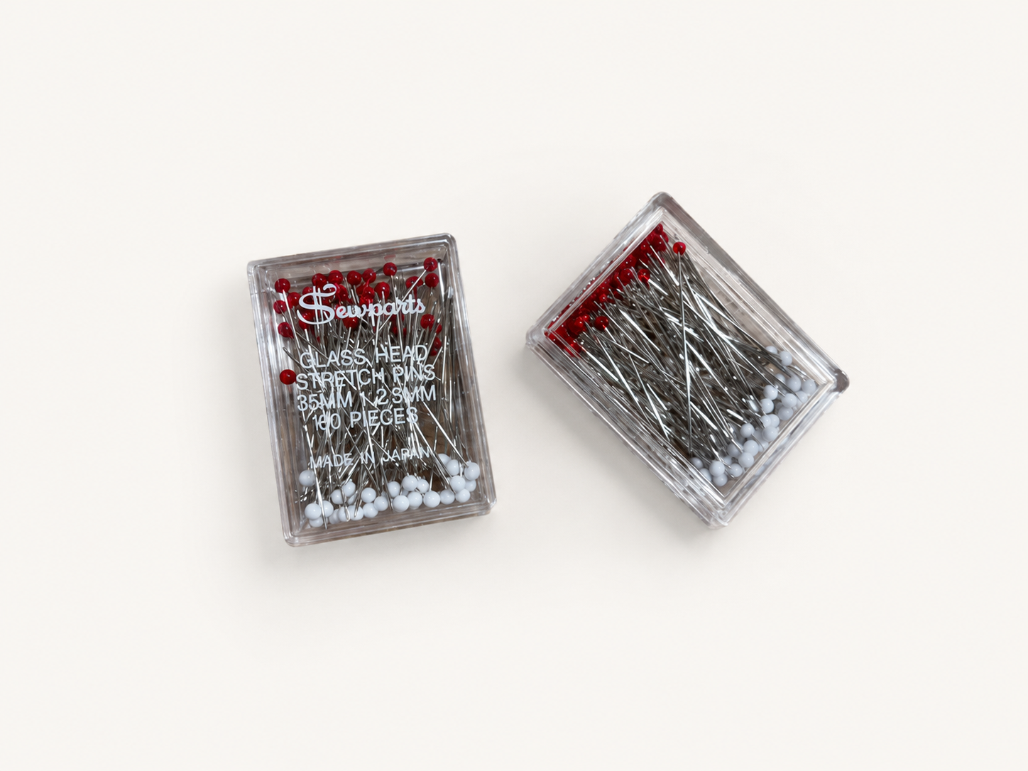 Two clear plastic cases of Sewparts glass head stretch dressmaking pins available at the RMIT Store, shown on a light grey studio background with visible white Sewparts branding and product details printed on the front case, containing silver metal pins with red and white rounded glass heads arranged inside transparent rectangular storage containers.