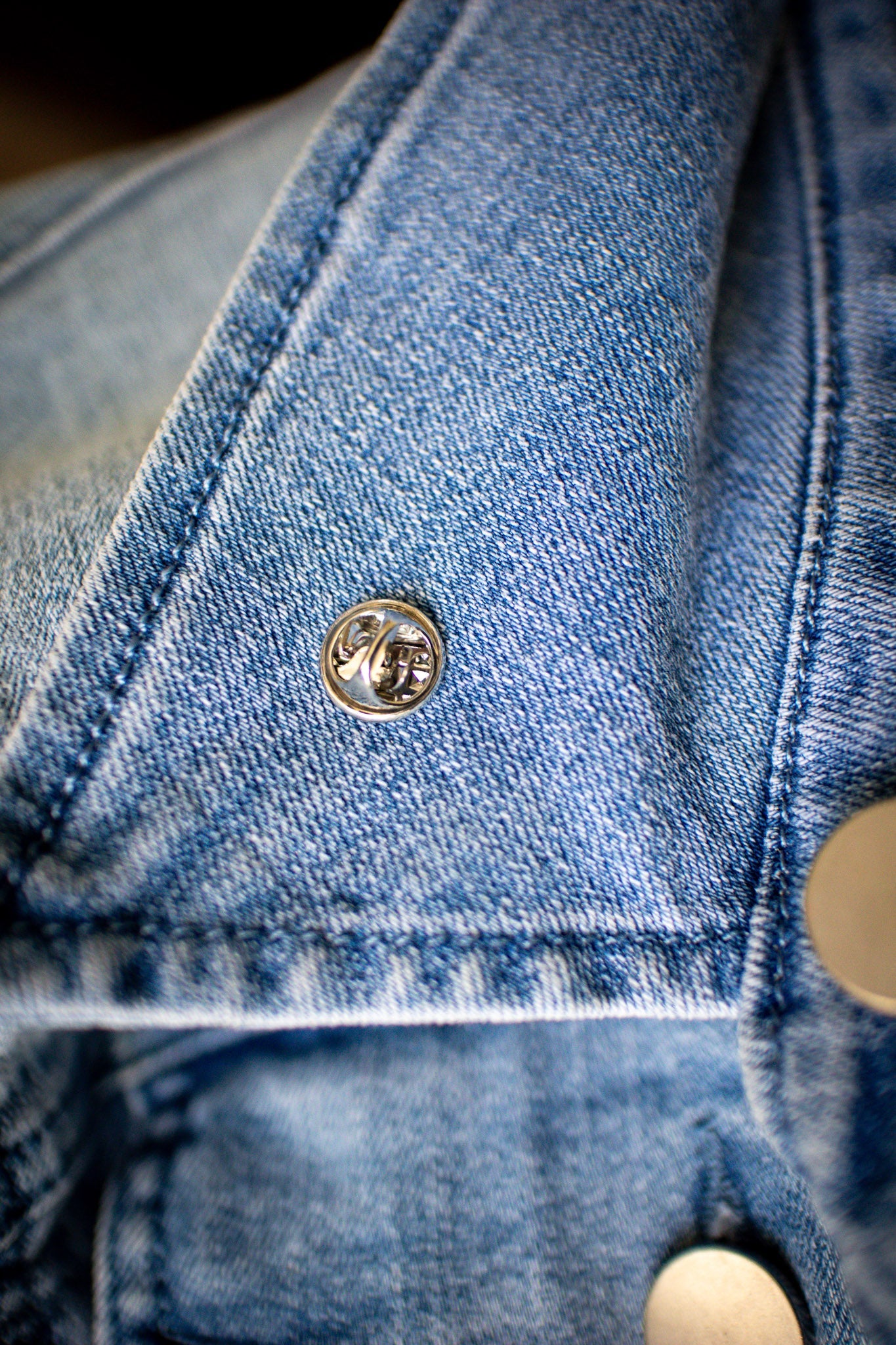 Close-up view of the back of an enamel pin secured to a blue denim collar. The pin’s butterfly clutch backing is visible, firmly fastened to the fabric. The texture of the denim and stitching detail of the collar are clearly shown. This image highlights the pin's attachment style and secure fit, available at the RMIT Store.