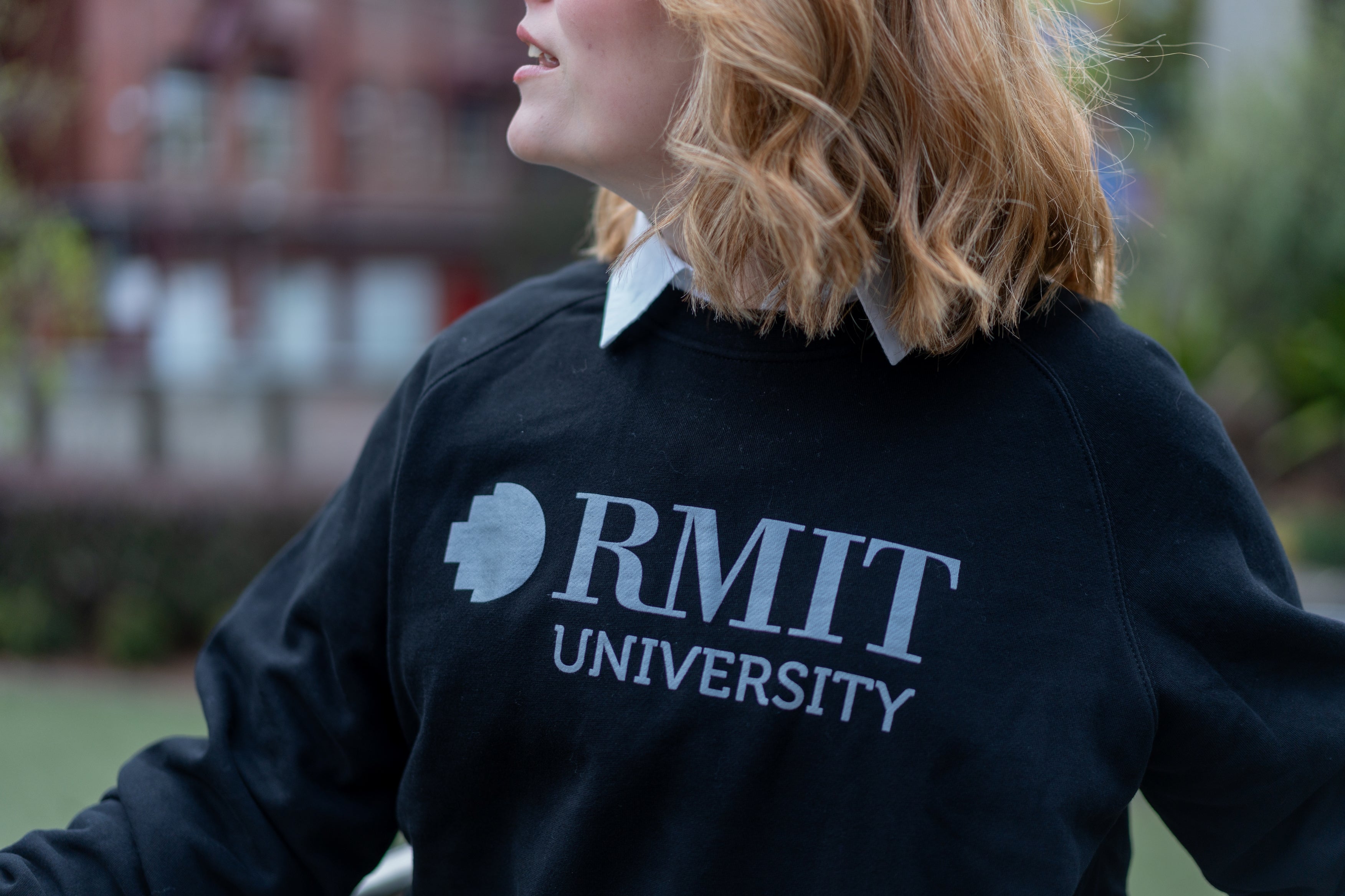 Close-up of a model in the black RMIT Essential Crew sweater, prominently displaying the white RMIT University logo. The sweater is worn over a white collared shirt, highlighting the logo's design. Part of the RMIT Store collection, this sweater is crafted for style and warmth.
