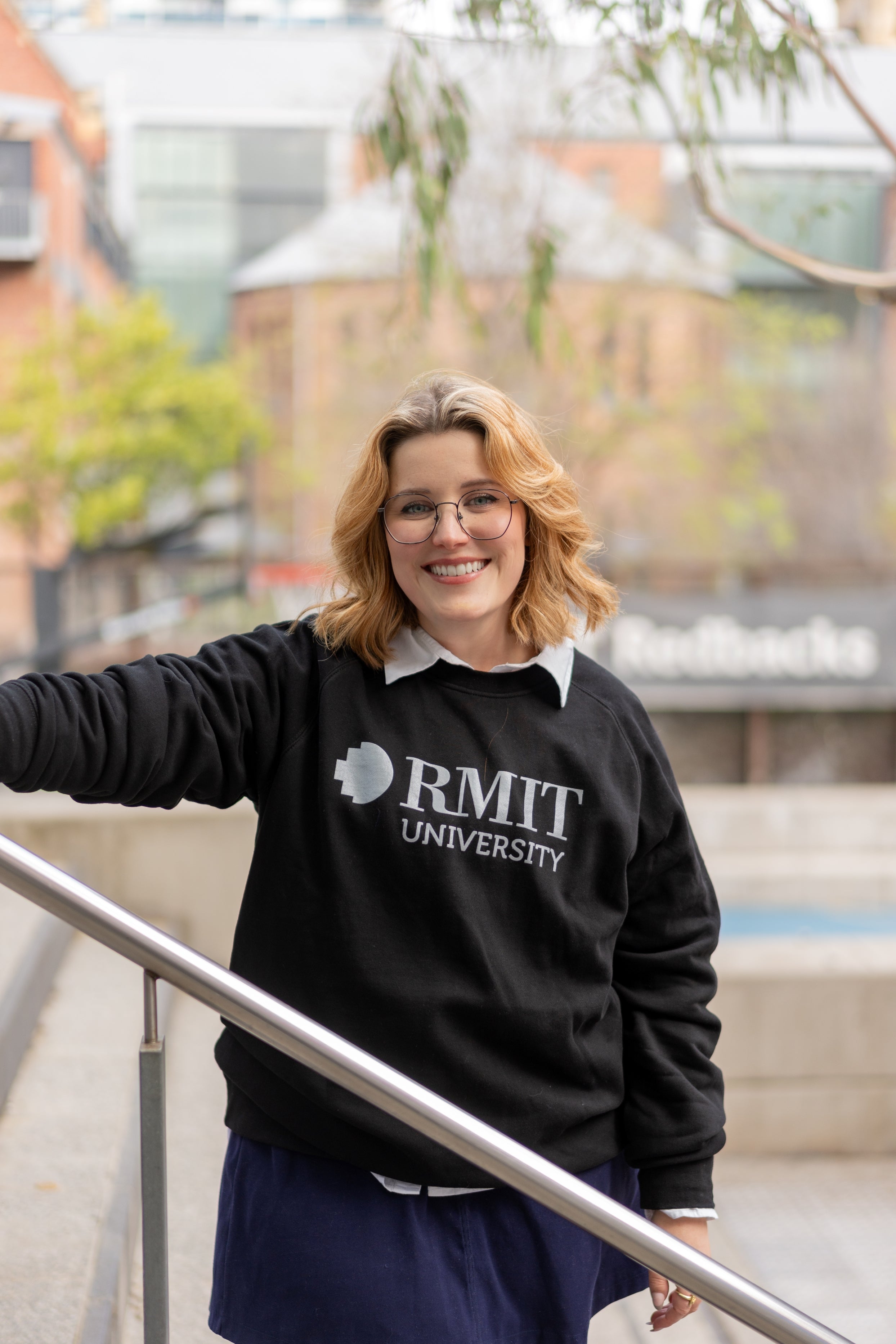 Model posing with a black RMIT Essential Crew sweater, featuring the RMIT University logo in white, captured on stairs in an urban environment. Styled over a white collared shirt, the sweater offers comfort and university pride, available at the RMIT Store.