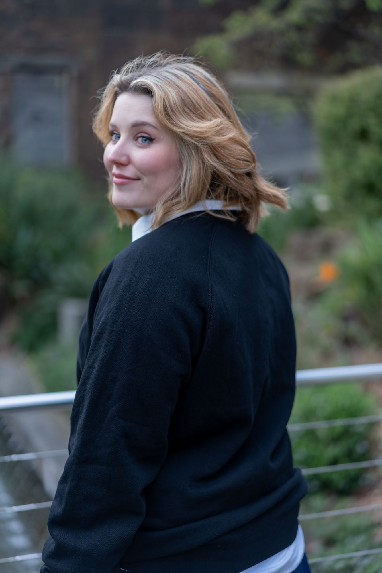 Model in a black RMIT Essential Crew sweater, with a subtle outdoor setting providing a natural backdrop. The sweater displays a clean, minimalistic style while layered over a white collared shirt. Part of the RMIT Store collection.
