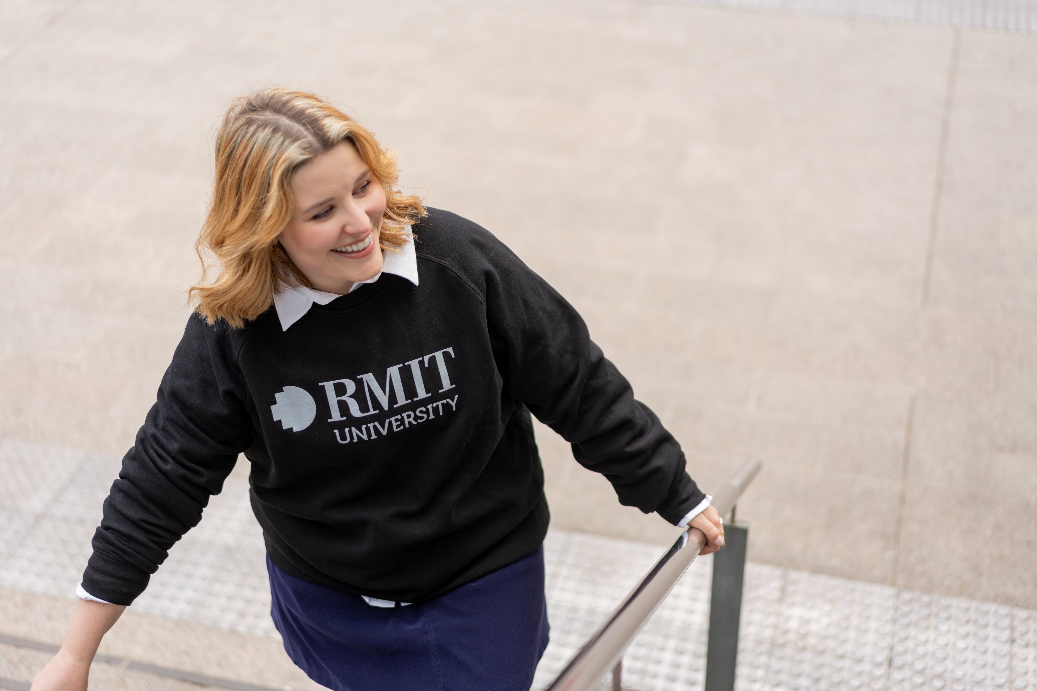Model wearing the RMIT Essential Crew sweater in black with a white RMIT University logo. The sweater is layered over a white collared shirt and featured while the model ascends a set of outdoor stairs. This versatile sweater from the RMIT Store blends style and comfort for everyday wear.