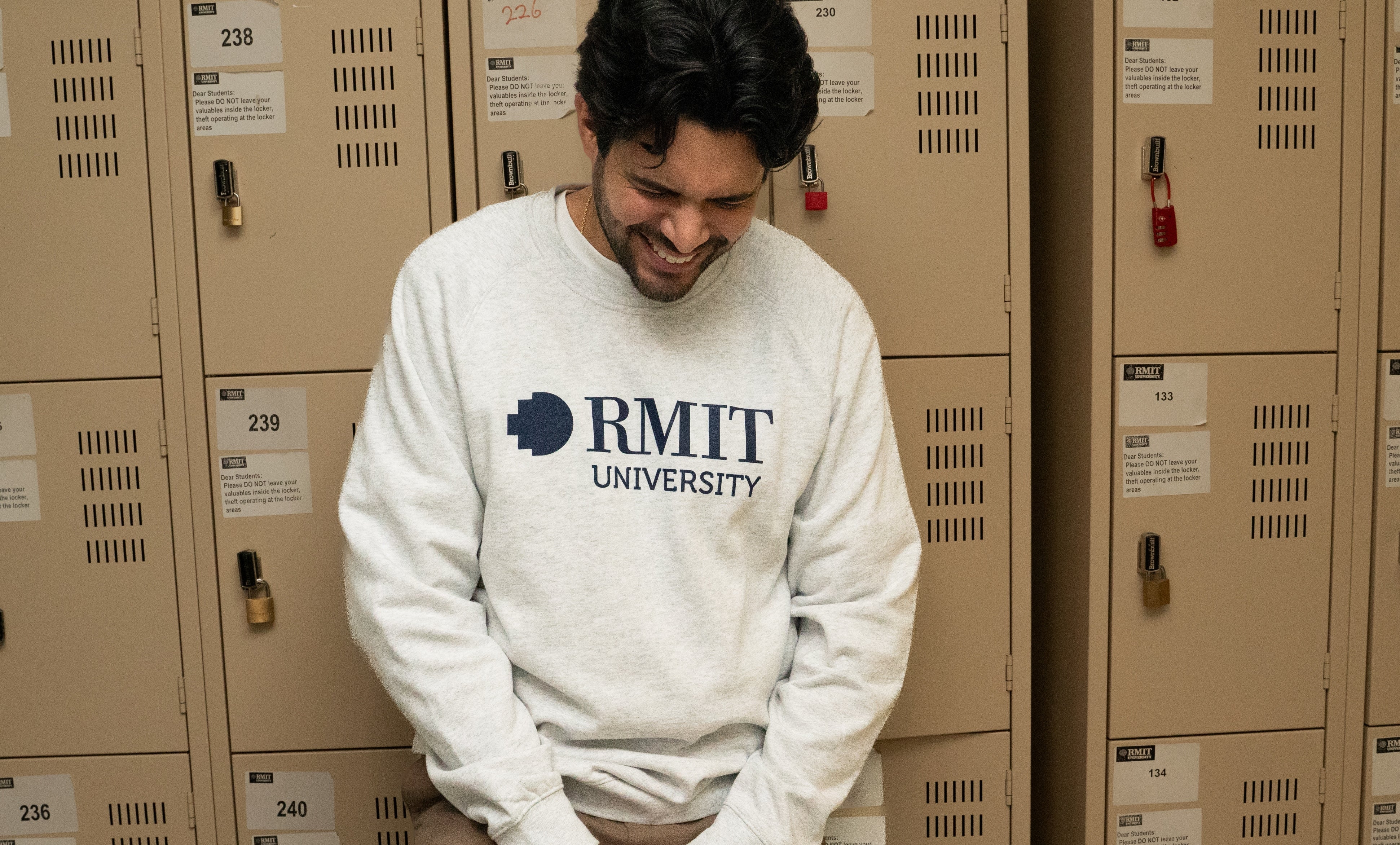 Model positioned in front of lockers wearing a white marle RMIT Essential Crew sweater, adorned with a navy RMIT University logo. The backdrop of lockers provides a collegiate atmosphere, emphasising the sweater's casual appeal. Available at the RMIT Store, perfect for students and alumni.