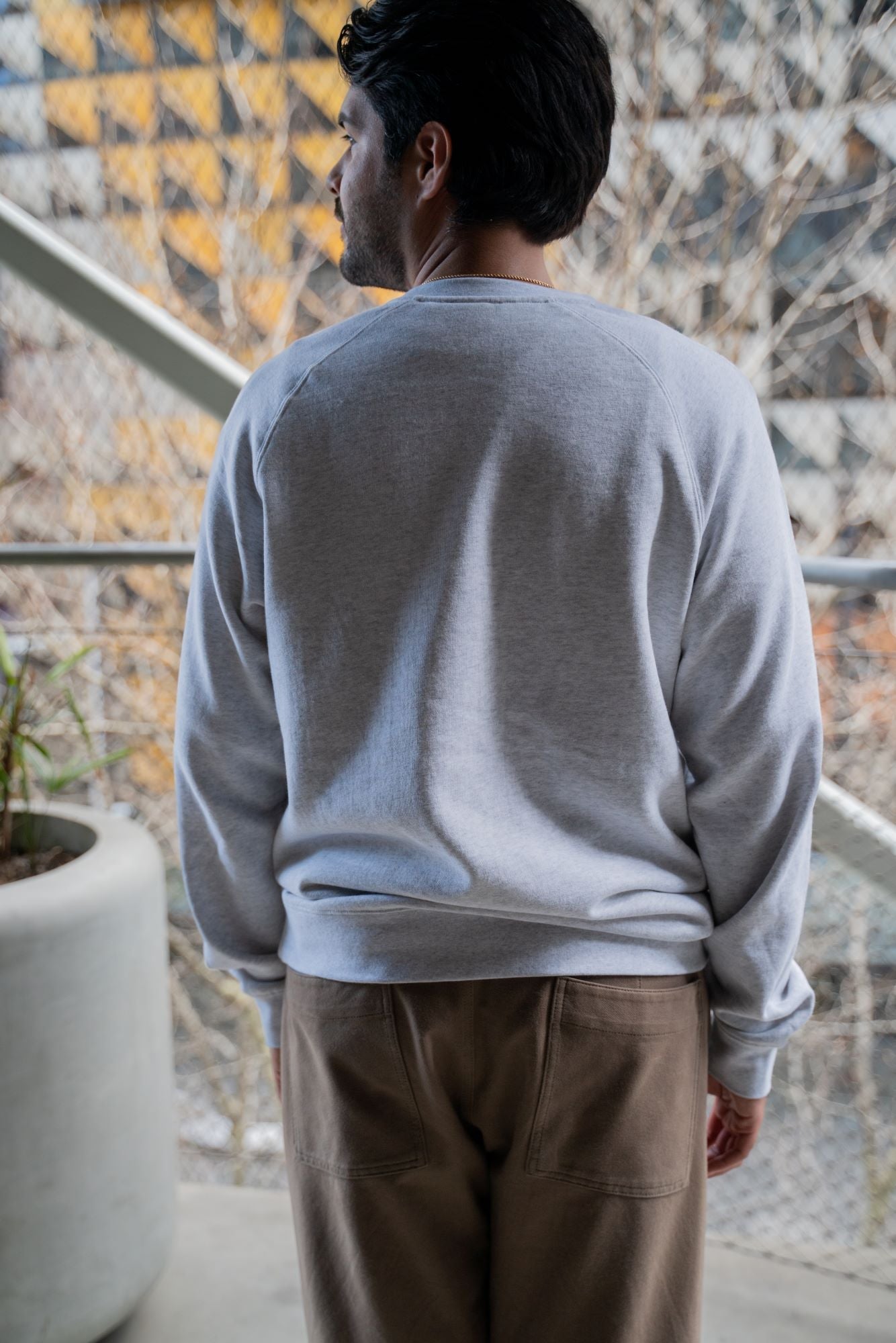 Back view of a model wearing the white marle RMIT Essential Crew sweater, showcasing its casual fit and design. The model is standing on a balcony with an urban backdrop, highlighting the sweater’s simplicity and comfort. Available at the RMIT Store.