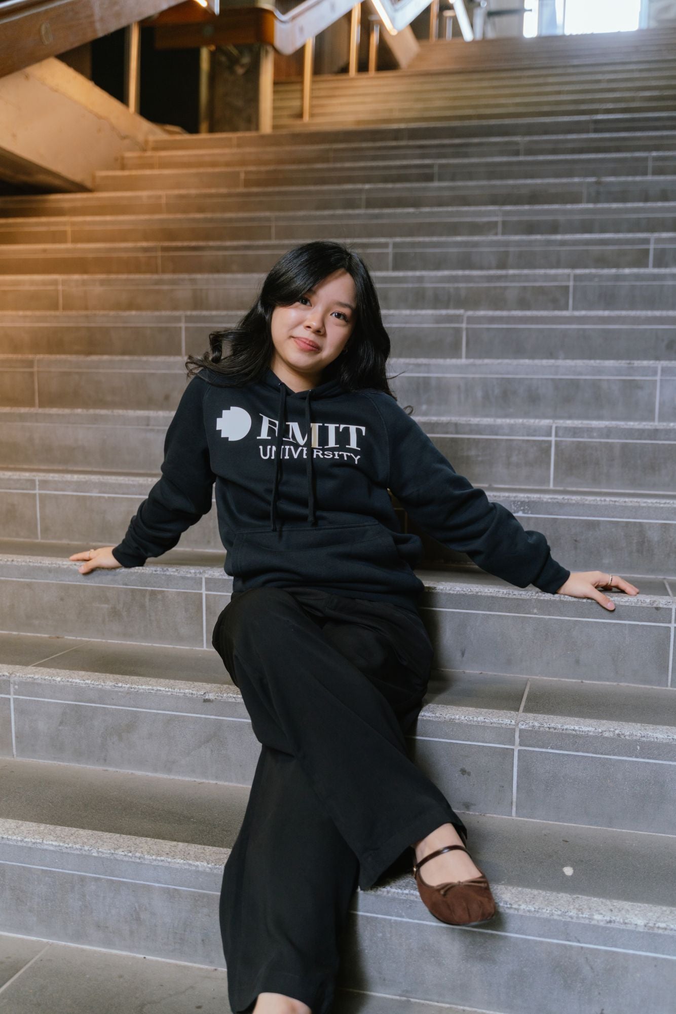 Model seated on wide indoor staircase wearing RMIT Essential Hoodie in navy with large white RMIT University logo across the chest, styled with black pants and brown flats, arms extended along steps in a relaxed pose within a modern stairwell environment, available at the RMIT Store.