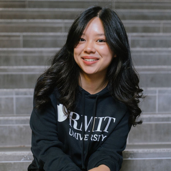 Model seated on indoor concrete stairs wearing RMIT Essential Hoodie in navy featuring a bold white RMIT University logo printed across the chest, with drawstring hood and long sleeves, paired with black pants, smiling towards camera in a softly lit stairwell setting, available at the RMIT Store.