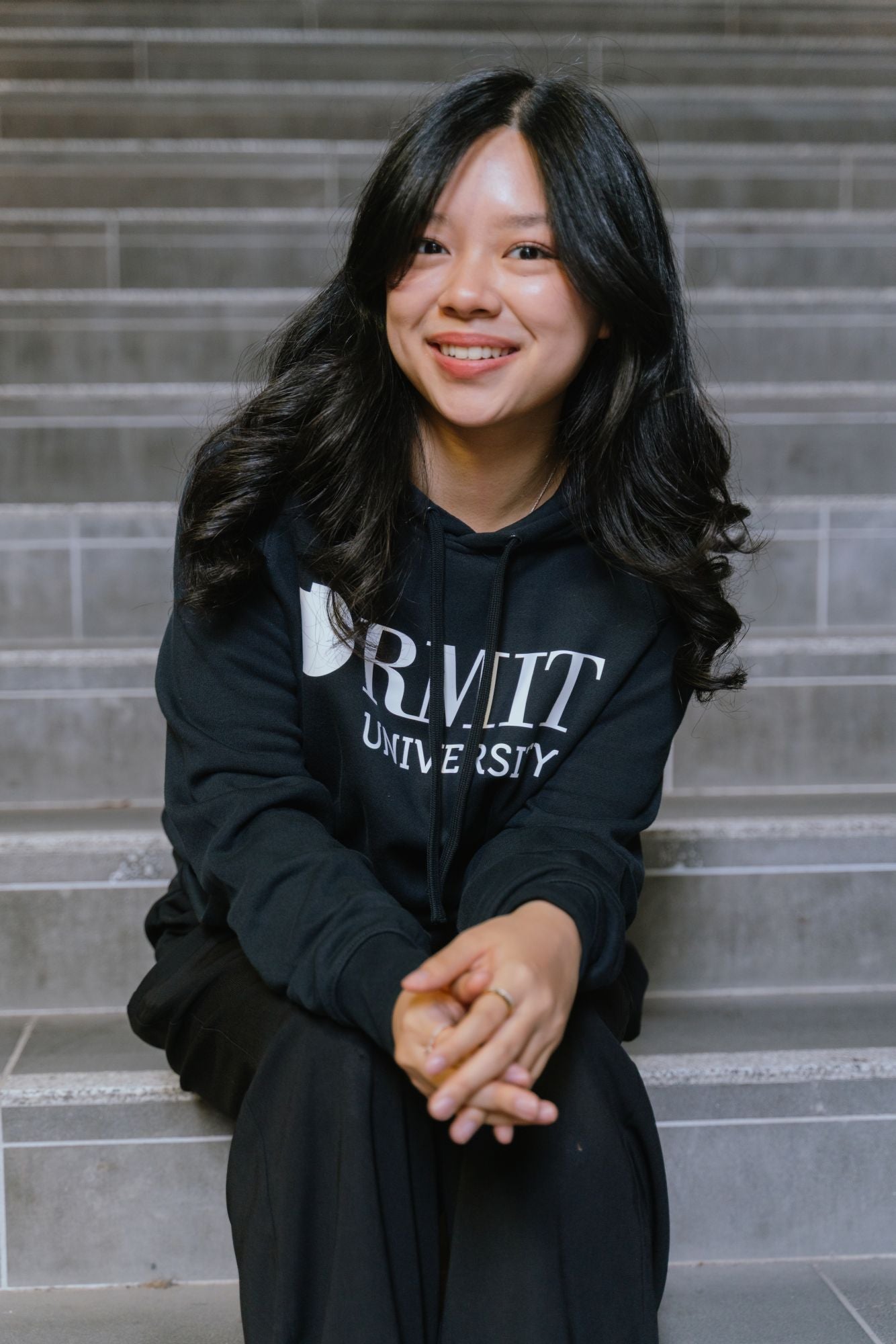 Model seated on indoor concrete stairs wearing RMIT Essential Hoodie in navy featuring a bold white RMIT University logo printed across the chest, with drawstring hood and long sleeves, paired with black pants, smiling towards camera in a softly lit stairwell setting, available at the RMIT Store.