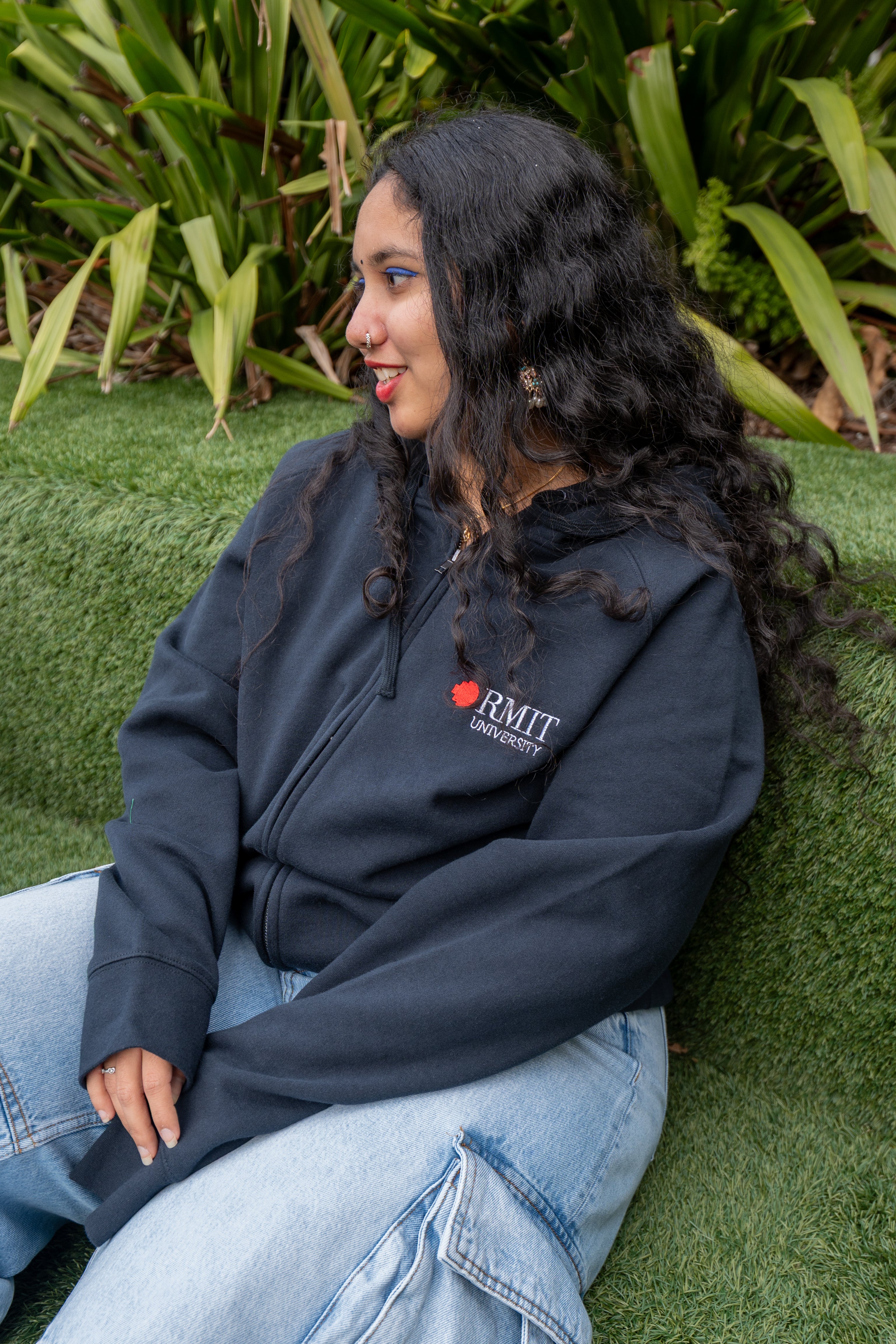 Close-up of a seated female model wearing the navy Essential Jacket on green grass with large leafy plants behind; the red and white embroidered RMIT University logo is clearly visible on the left chest. She is showing the left side of her face. Available at the RMIT Store.