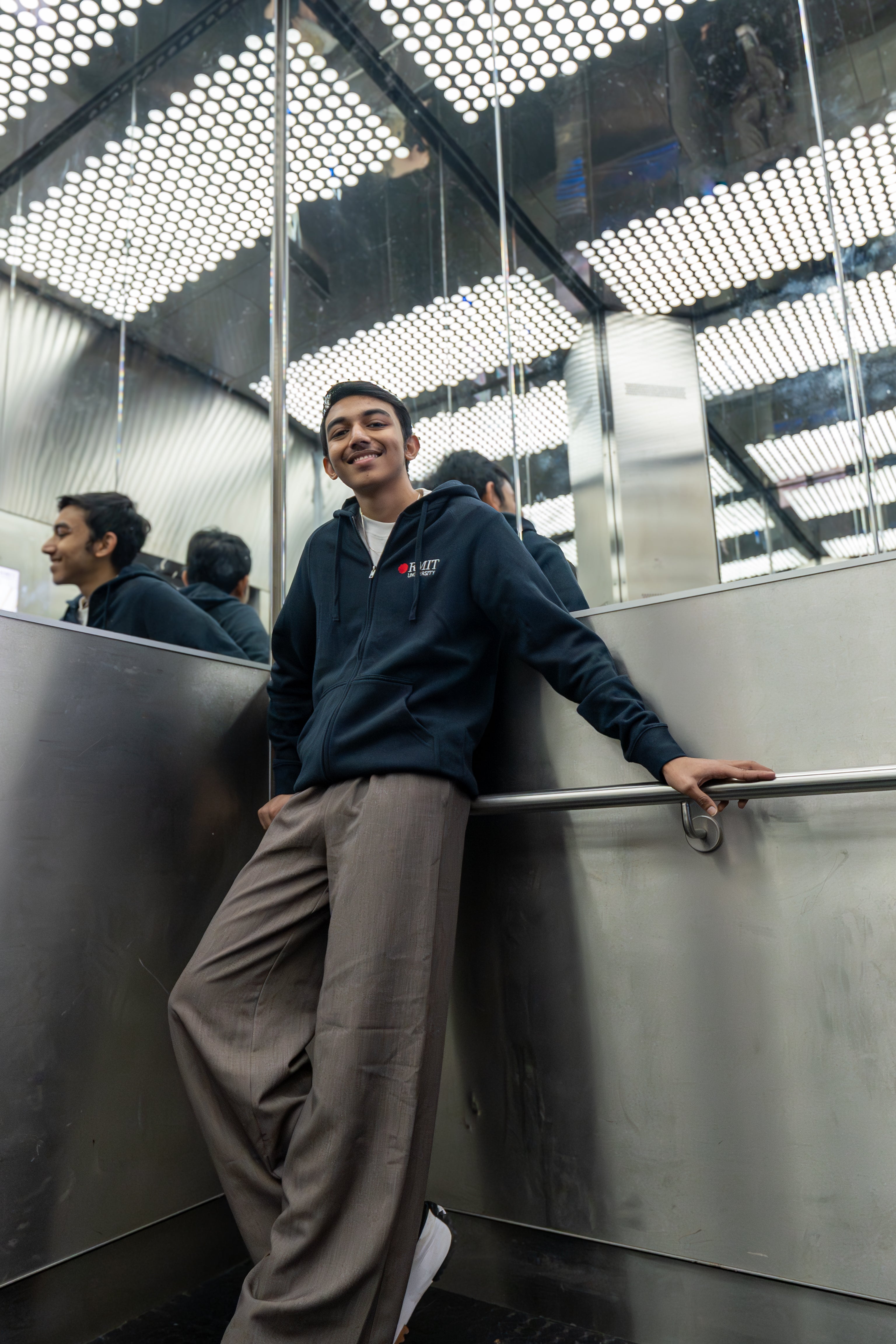 A male model wearing the navy Essential Jacket in a mirrored lift, smiling and leaning casually against the stainless steel interior; embroidered red and white RMIT University logo clearly visible on the left chest of the jacket. Available at the RMIT Store.