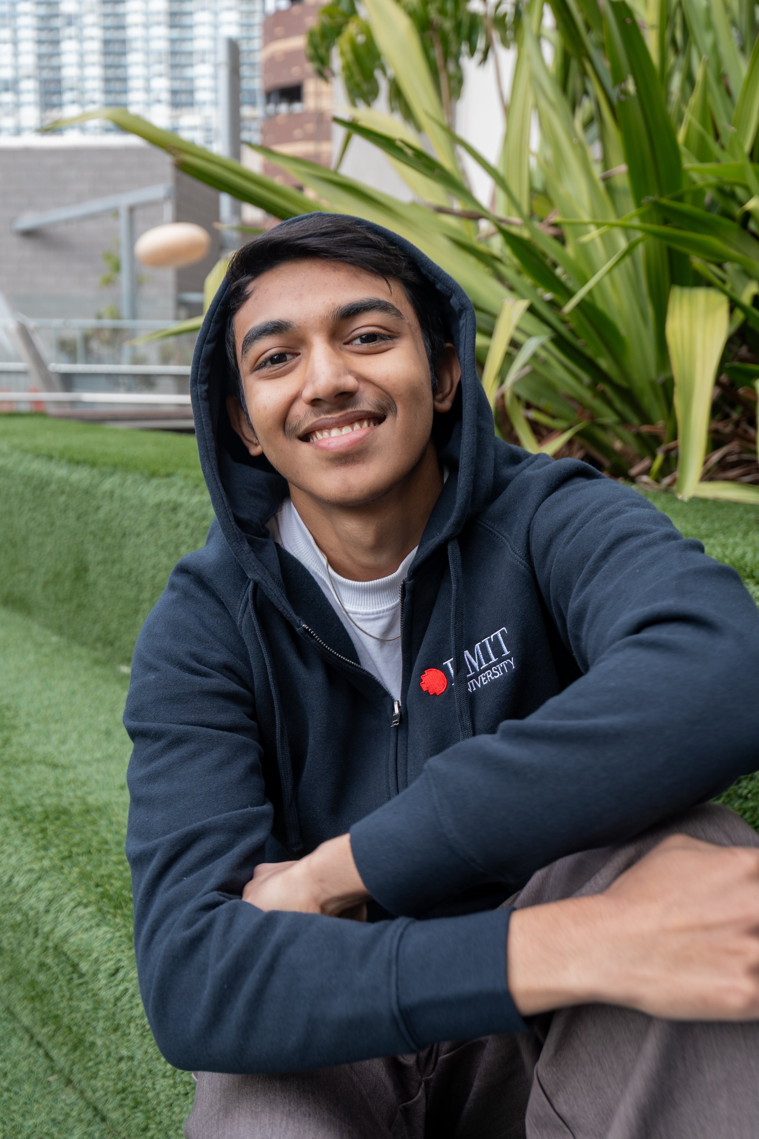 Close-up of a seated male model wearing the navy Essential Jacket on green grass with large leafy plants behind; the red and white embroidered RMIT University logo is clearly visible on the left chest. He is facing the camera. Available at the RMIT Store.