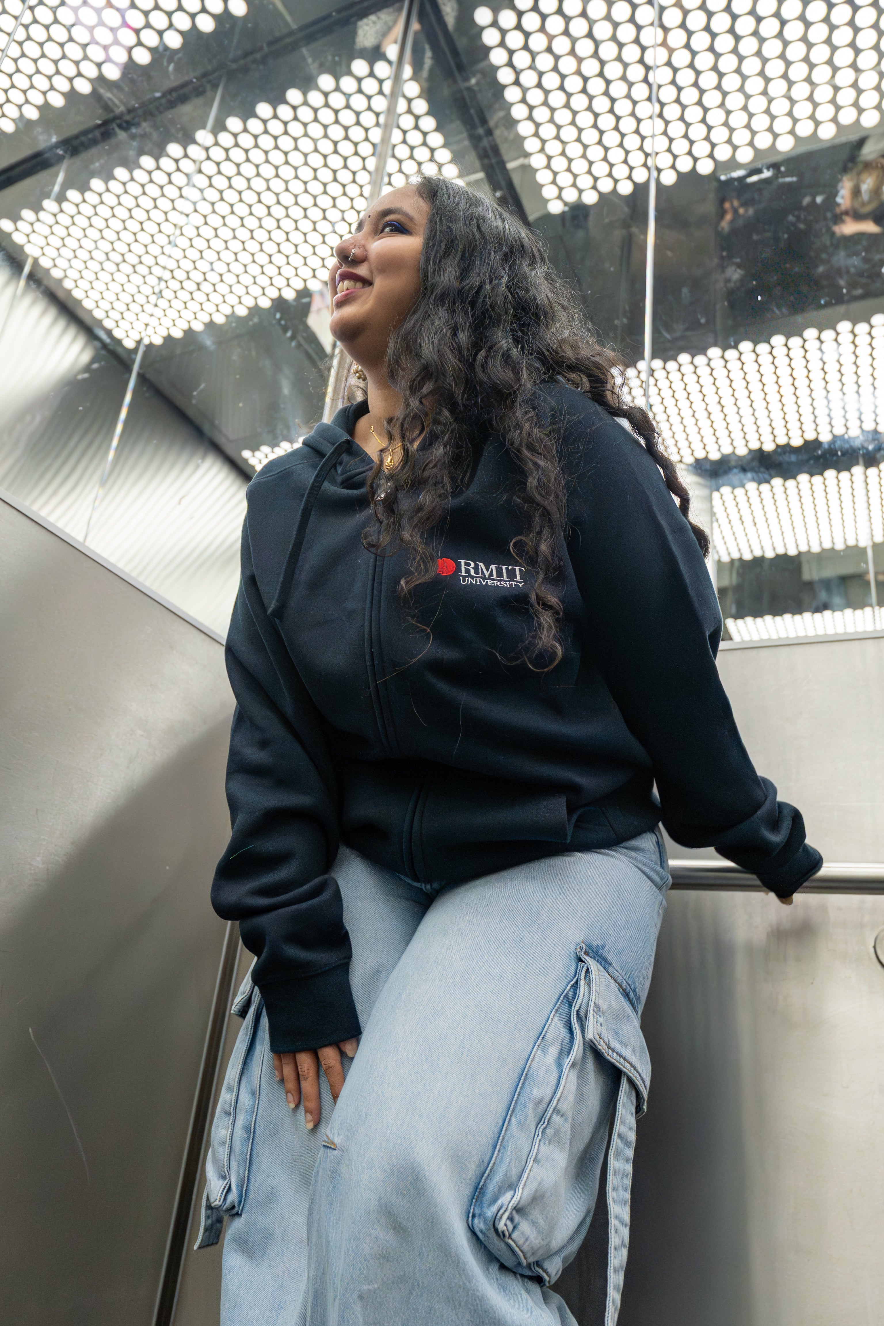 Two models wearing the navy Essential Jacket in a mirrored lift, both smiling and leaning casually against the stainless steel interior; embroidered red and white RMIT University logo clearly visible on the left chest of each jacket. Available at the RMIT Store.