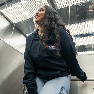 Two models wearing the navy Essential Jacket in a mirrored lift, both smiling and leaning casually against the stainless steel interior; embroidered red and white RMIT University logo clearly visible on the left chest of each jacket. Available at the RMIT Store.