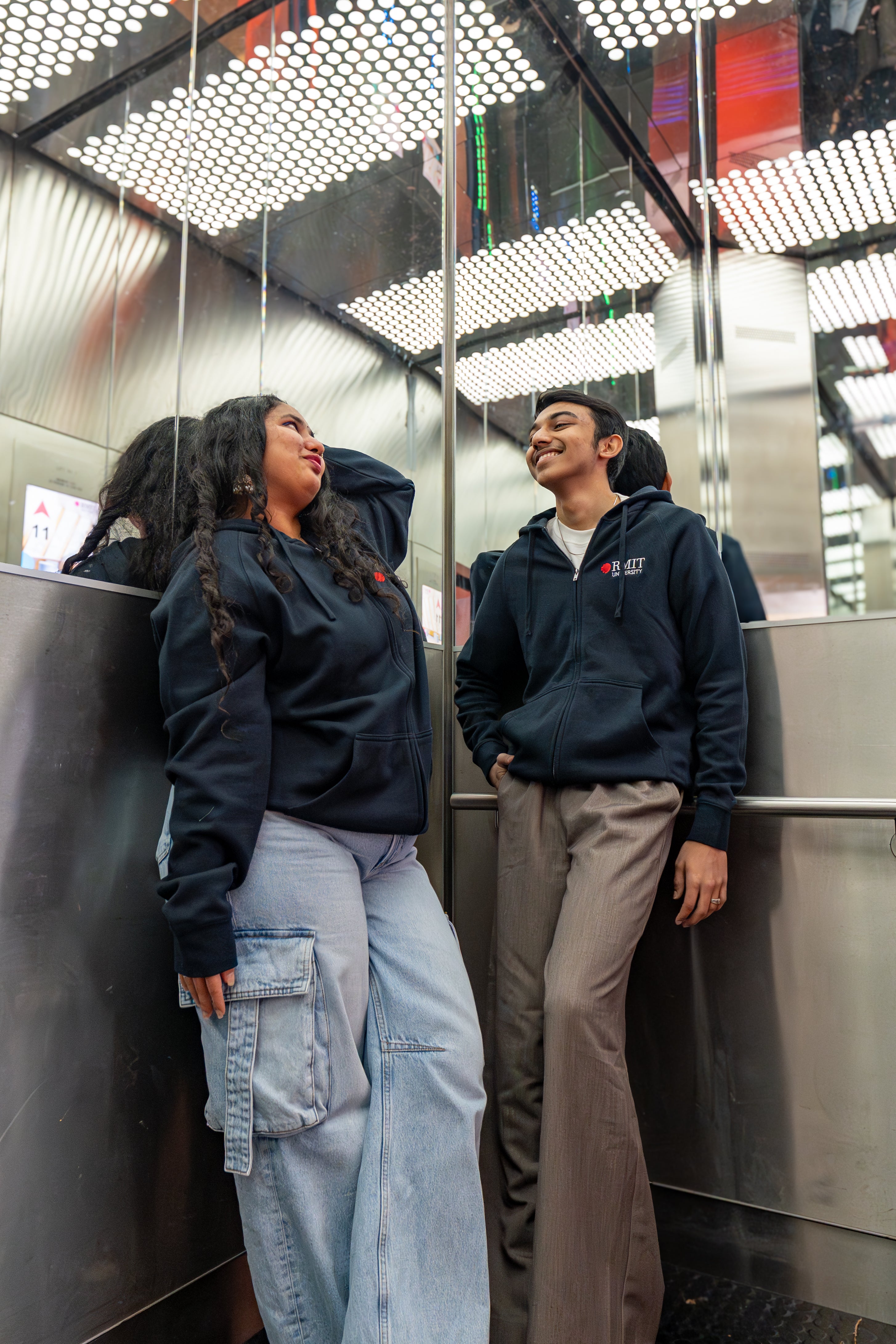 Two models wearing the navy Essential Jacket in a mirrored lift, both smiling and leaning casually against the stainless steel interior; embroidered red and white RMIT University logo clearly visible on the left chest of each jacket. Available at the RMIT Store.