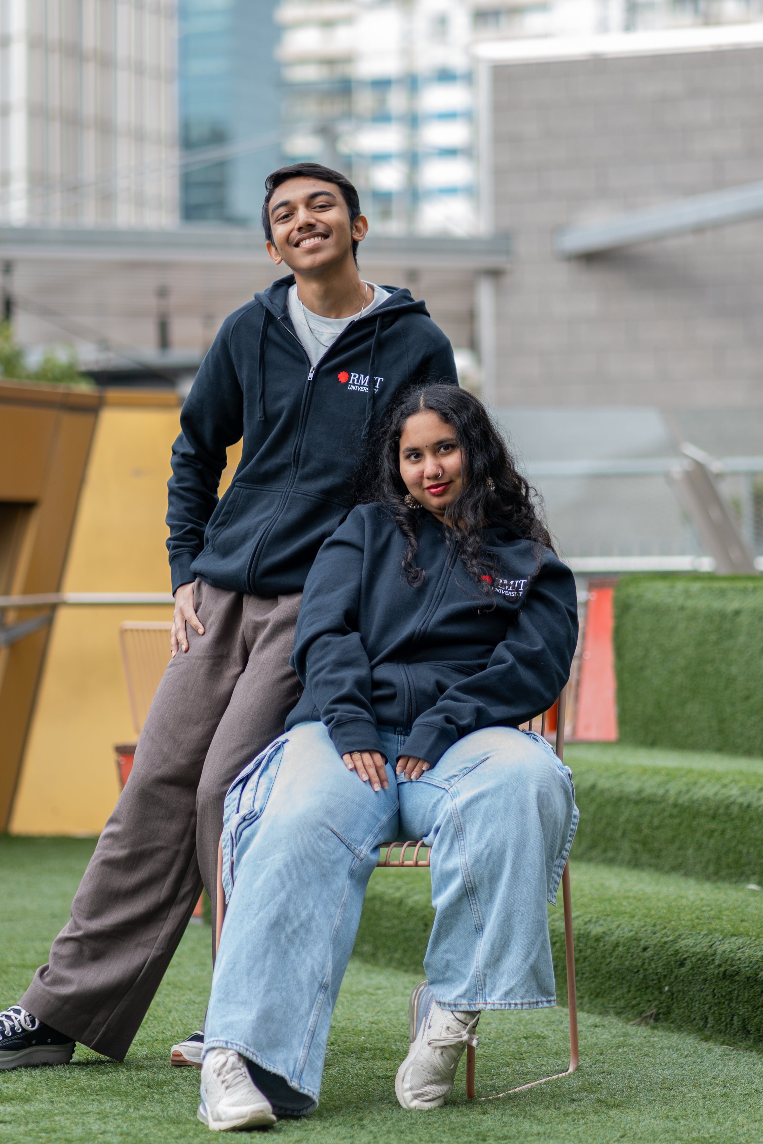 Two models wearing the navy Essential Jacket outdoors on green turf, one seated and one standing behind; both jackets feature the embroidered RMIT University logo in red and white on the left chest. Urban buildings visible in the background. Available at the RMIT Store.