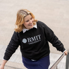 Model wearing the RMIT Essential Jumper in black with a white RMIT University logo. The jumper is layered over a white collared shirt and featured while the model ascends a set of outdoor stairs. This versatile sweater from the RMIT Store blends style and comfort for everyday wear.
