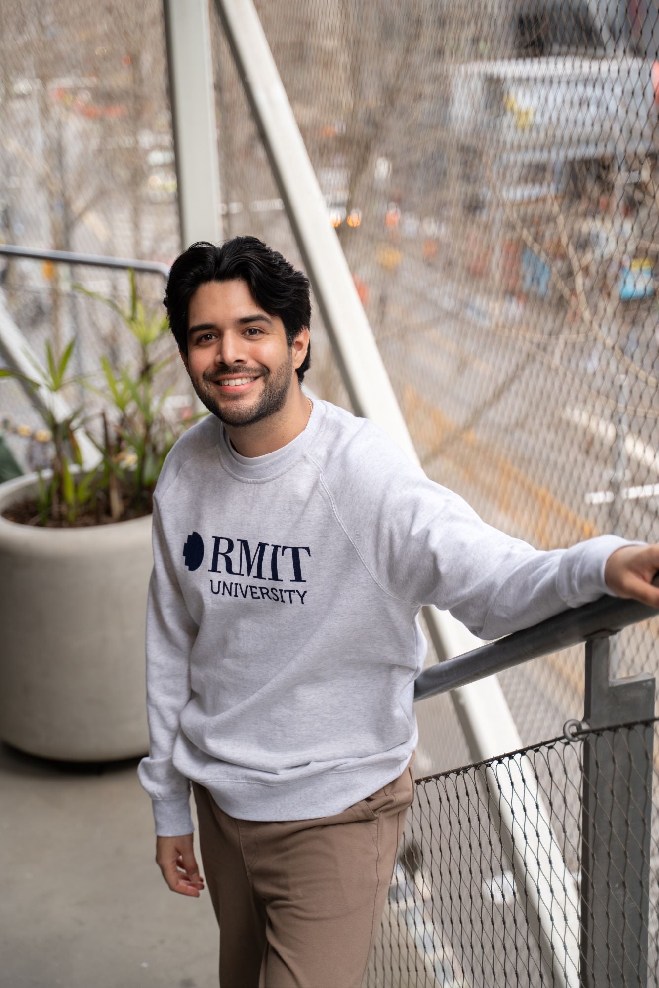 Model dressed in a white marle RMIT Essential Jumper featuring a navy RMIT University logo. The sweater is complemented by beige trousers, capturing a relaxed and sophisticated look. Photographed on a balcony setting, this apparel from the RMIT Store is ideal for showcasing university pride.